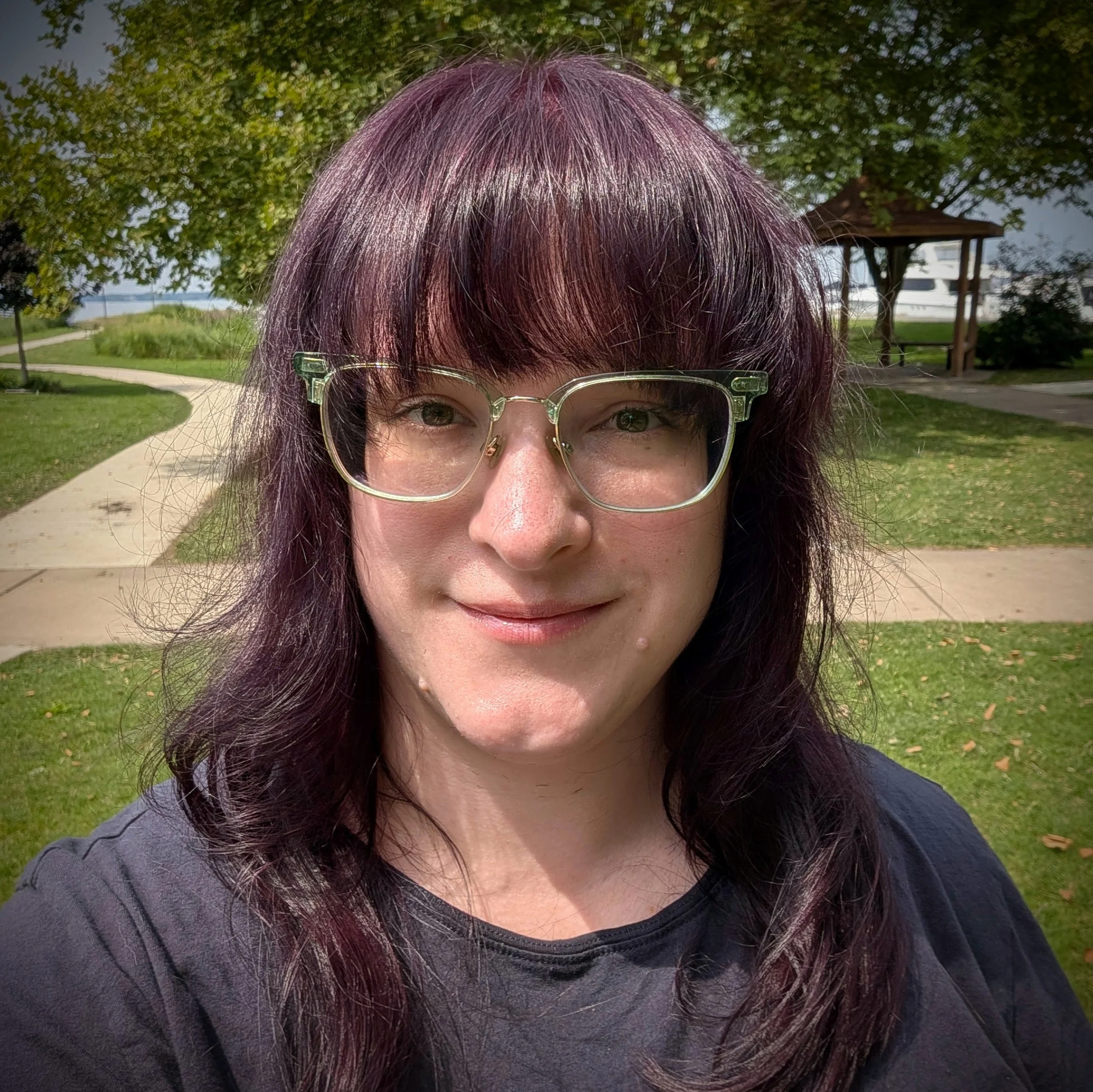 Close-up of a woman's face with glasses and purple hair, outdoors in a park with green trees and a pathway in the background.