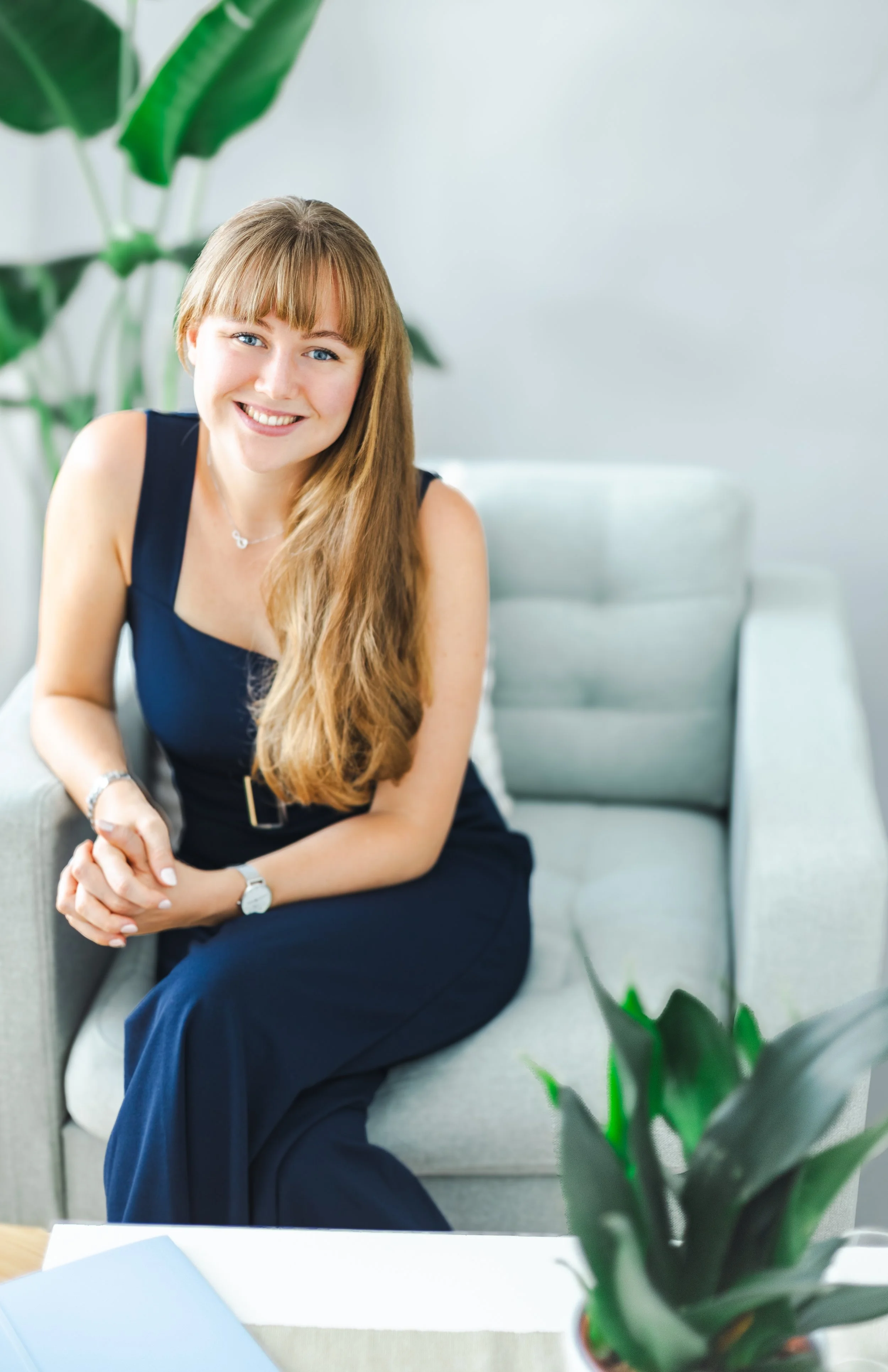 Ein Lächelnder junger Frau sitzt auf einem grauen Sofa, trägt ein dunkelblaues Kleid und eine Uhr, im Hintergrund sind Grünpflanzen und eine weiße Wand.