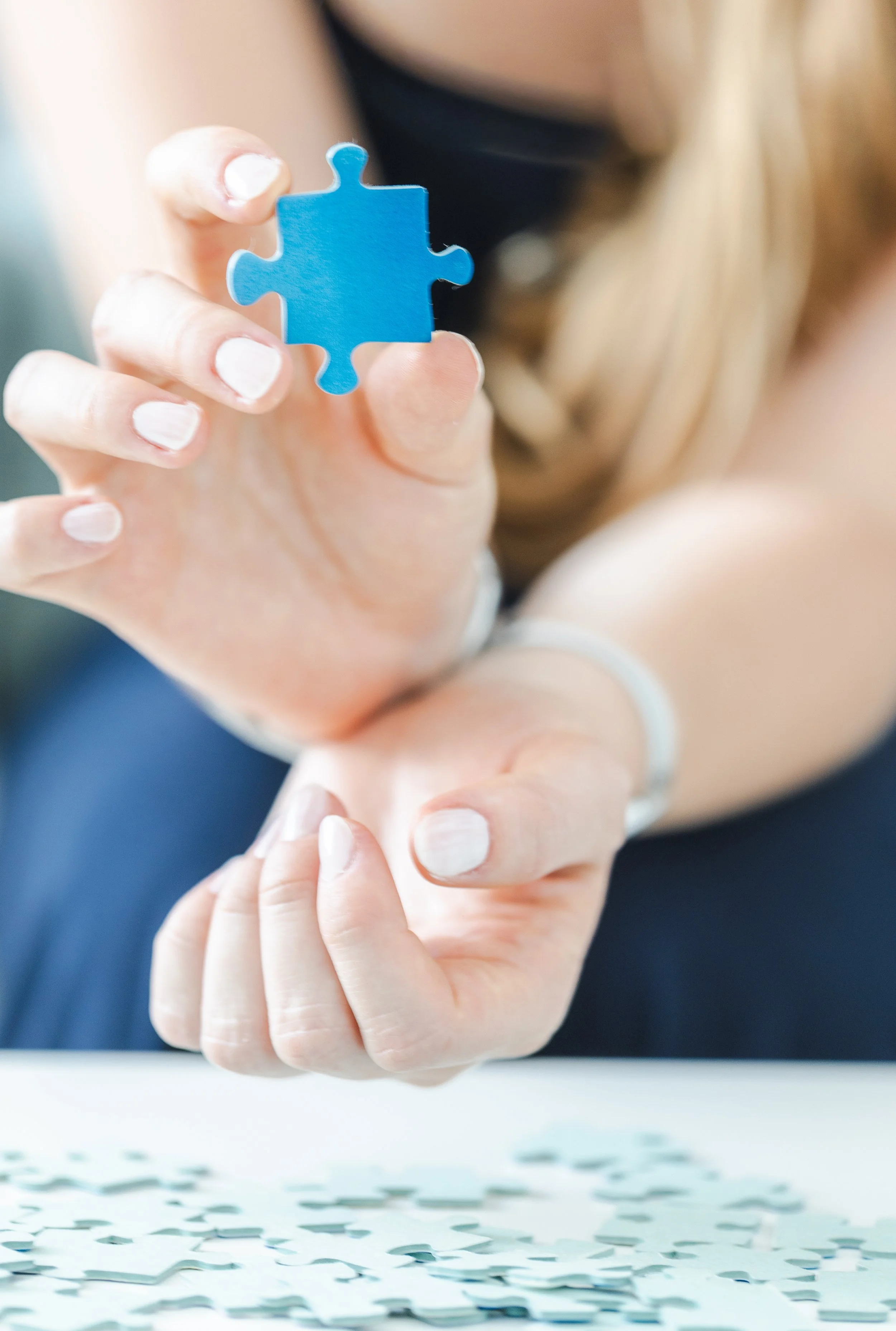 Eine Hand hält ein blaues Puzzlestück, während die andere Hand ein Puzzlestück auf einem Tisch mit mehreren Puzzlestücken hält.