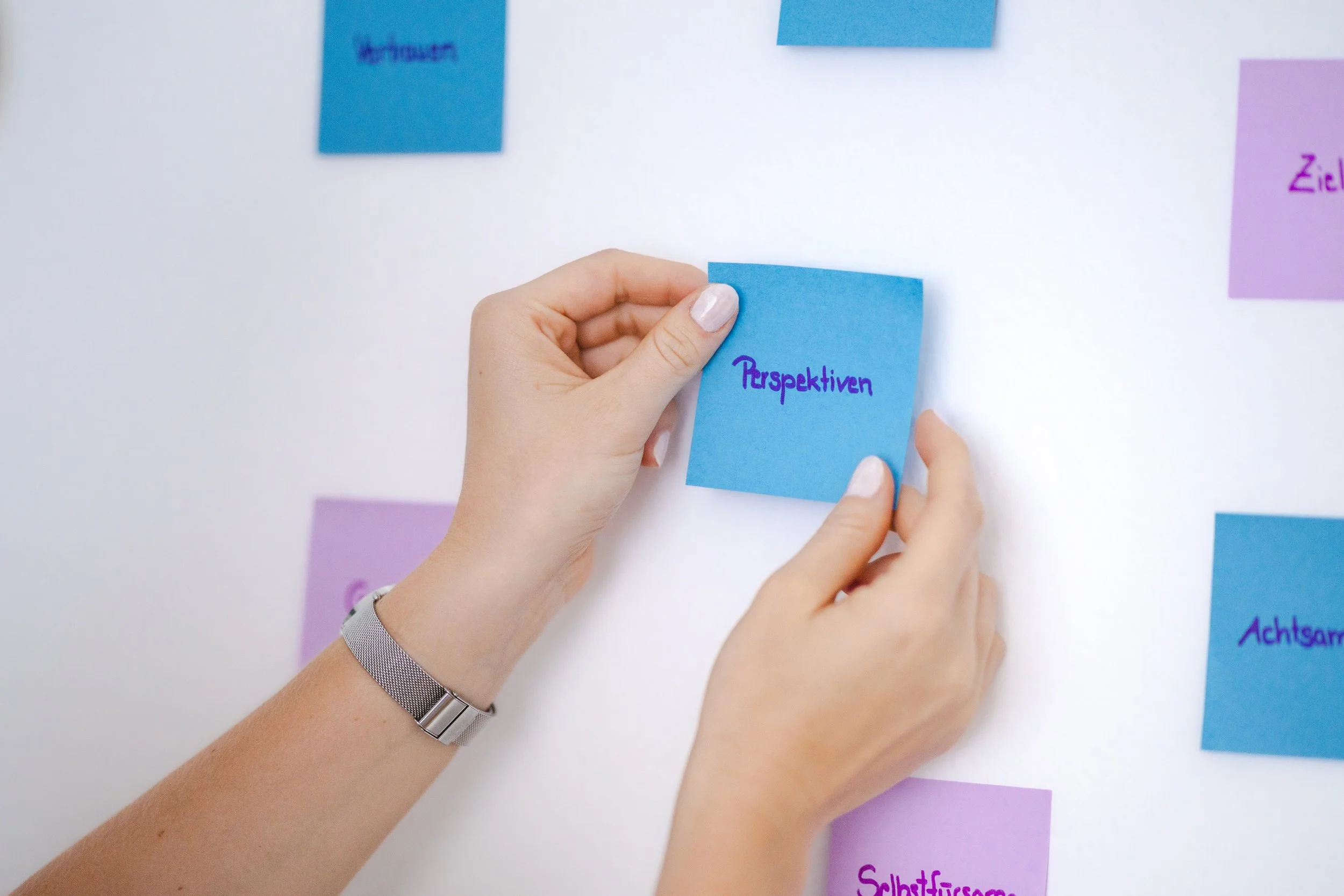 Hand legt einen blauen Notizzettel mit der Aufschrift 'Perspektiven' auf eine weiße Wand, umgeben von weiteren bunten Post-its.