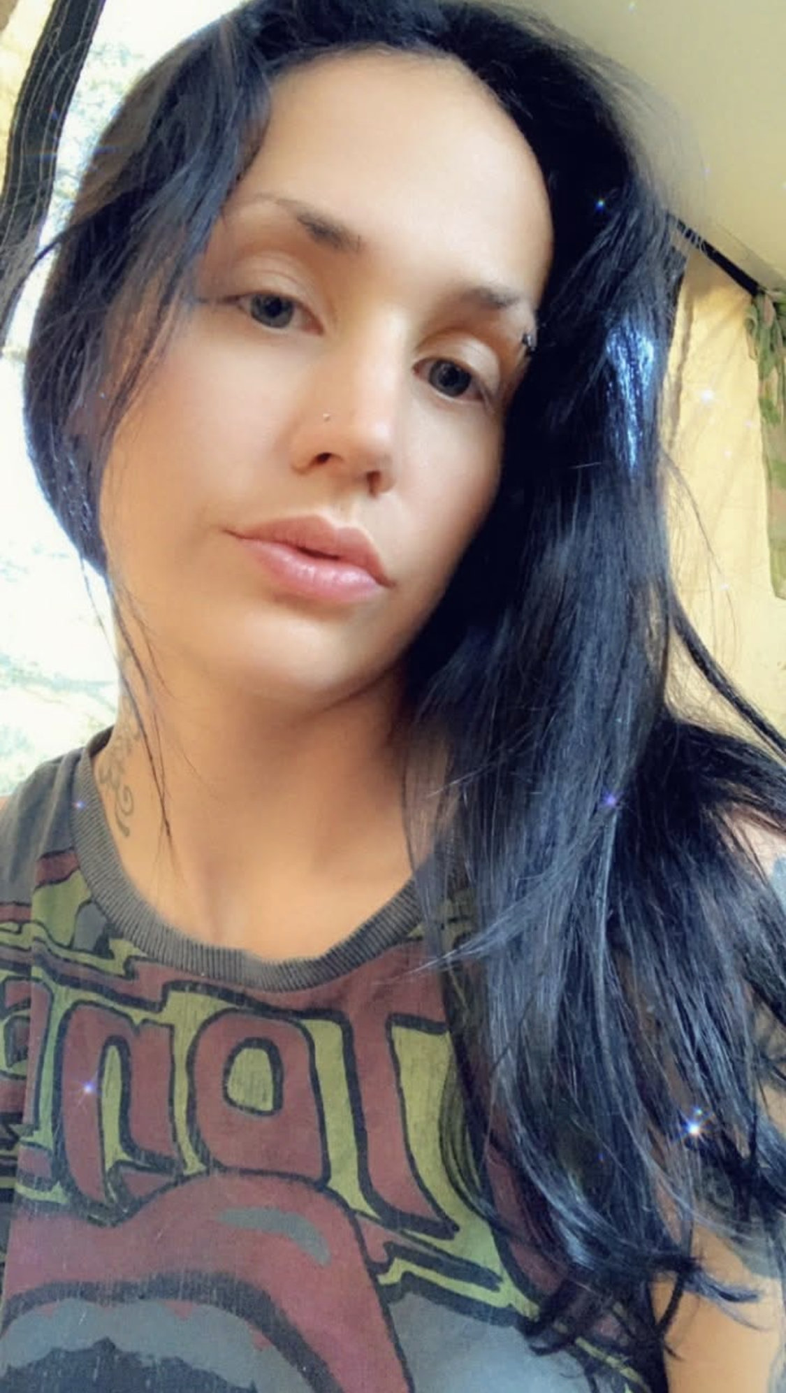Close-up of a woman with long black hair, light skin, blue eyes, and a septum piercing, wearing a graphic T-shirt with a colorful design.
