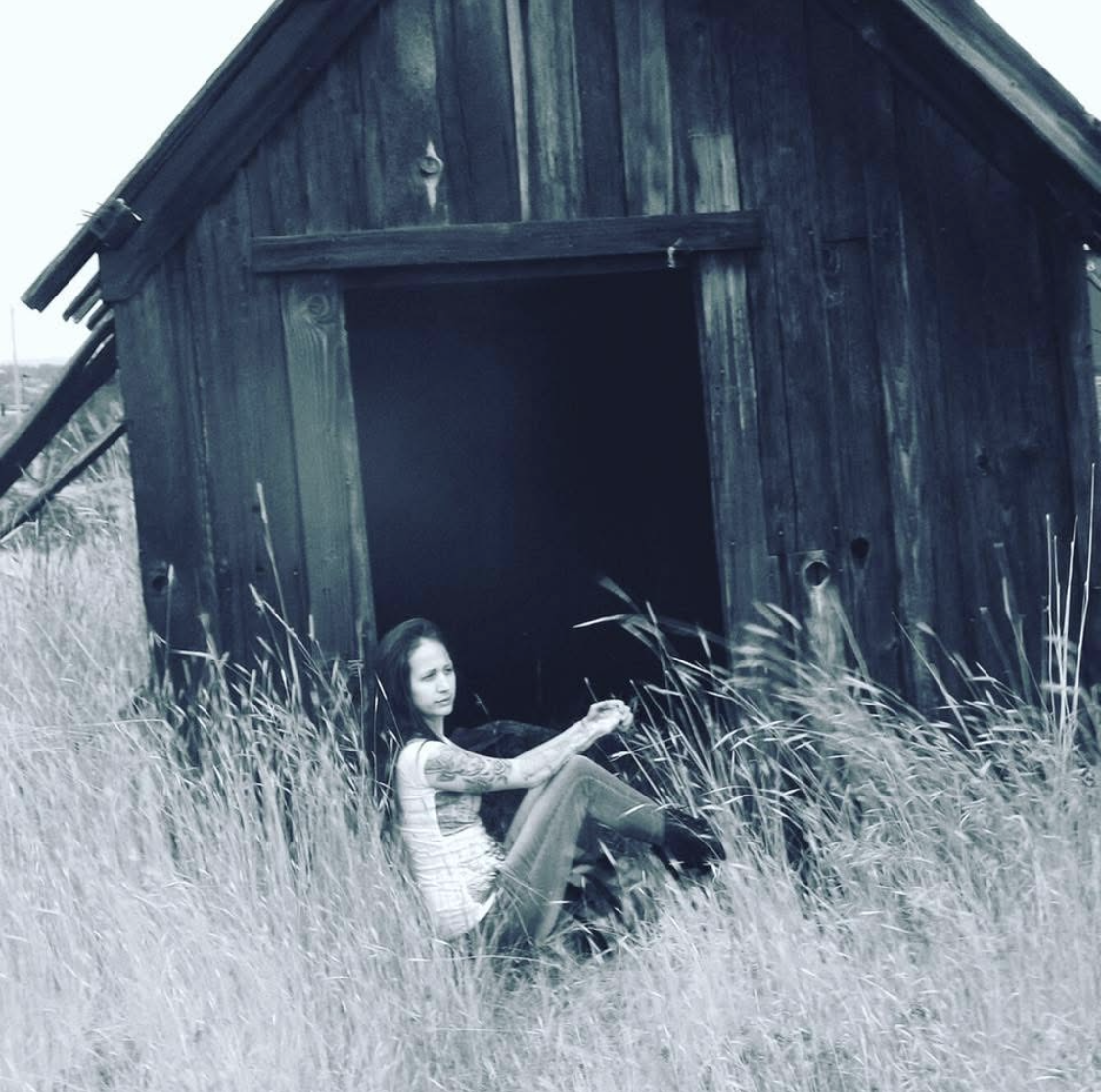 A woman with tattoos sitting outside a small, weathered wooden shed amongst tall grass, holding a cigarette.