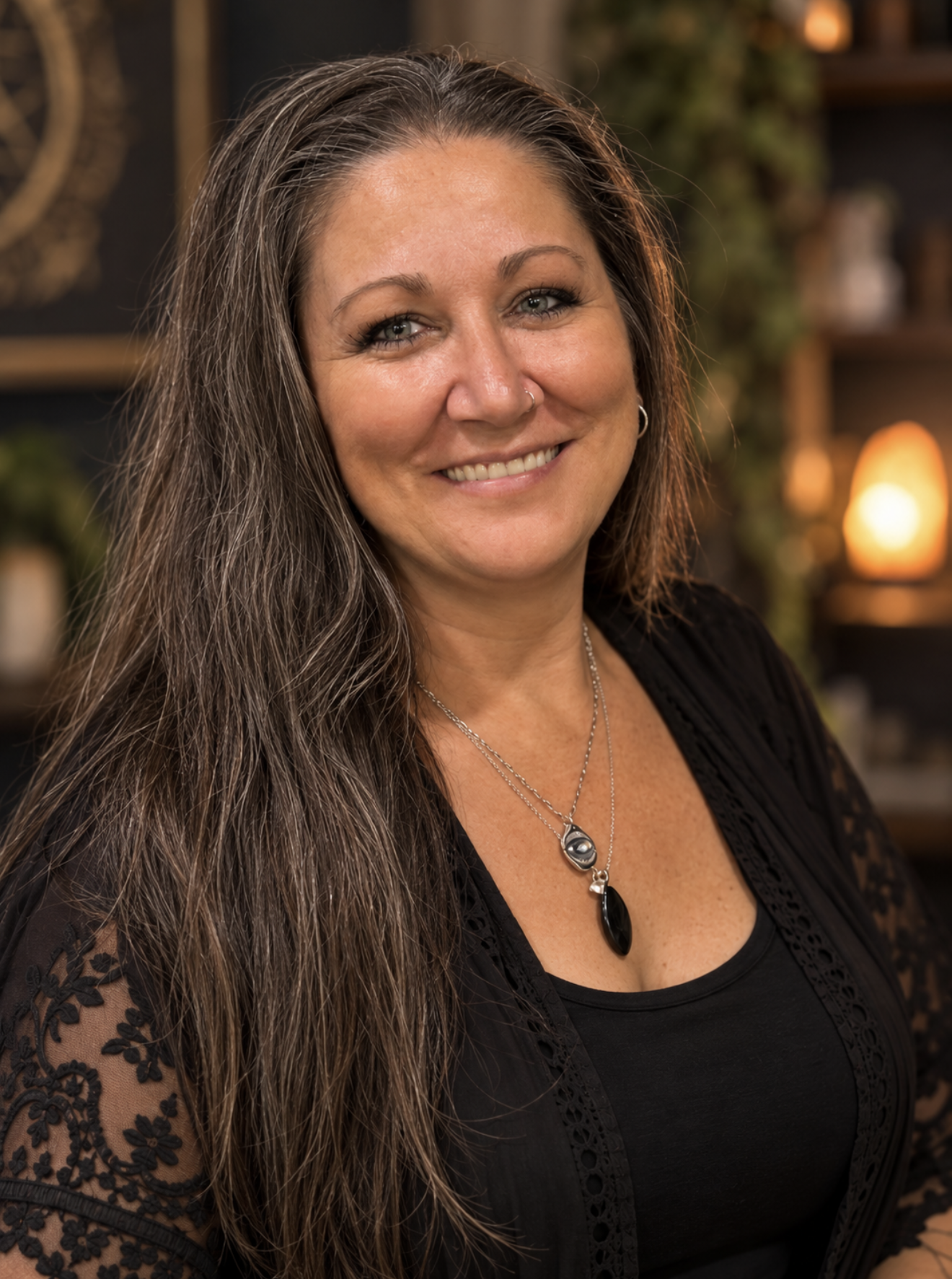 A smiling woman with long gray hair, wearing a black top with lace details and silver jewelry, in a warmly lit indoor setting.