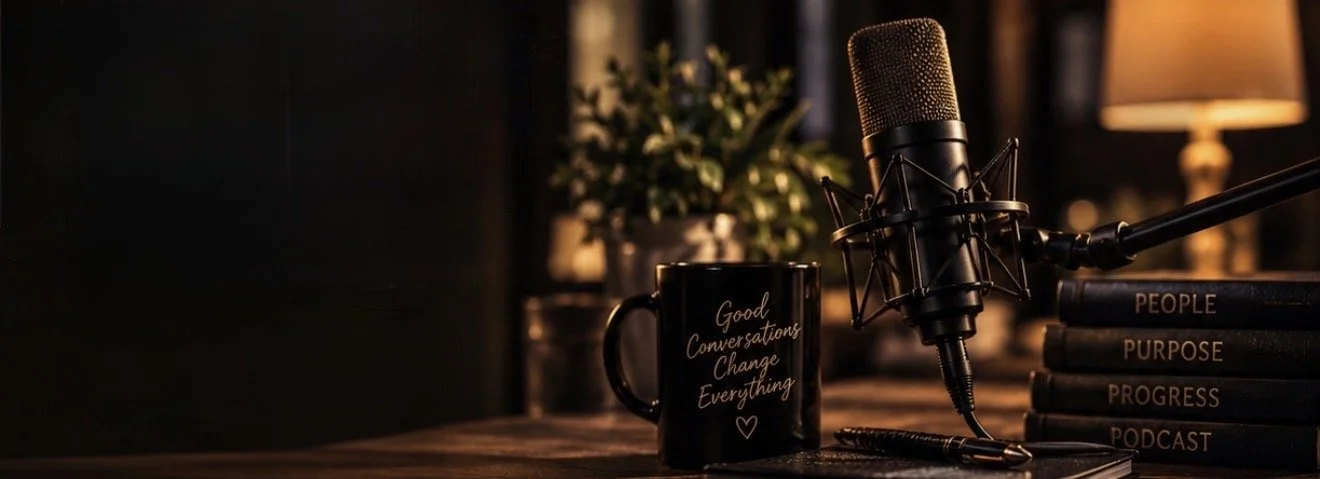 A microphone, a coffee mug with the text "Good Conversations Change Everything," a stack of books, and a lamp on a wooden table in a cozy, dimly lit room.