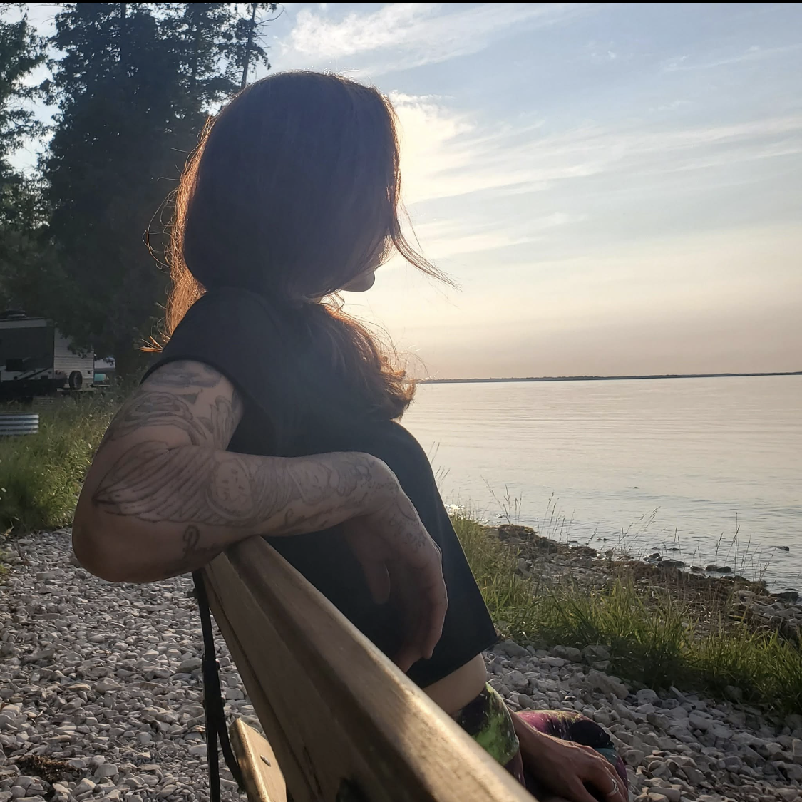A person sitting on a park bench near a body of water during sunset, with tattoos on their arm and long hair, gazing at the horizon.