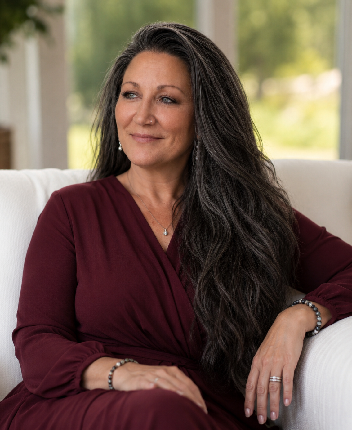 A woman with long dark hair wearing a maroon dress and jewelry, sitting on a white couch in a bright room with windows showing green trees outside.
