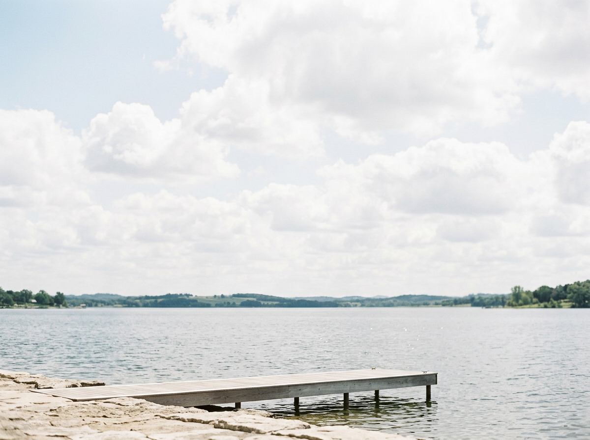 A wooden dock extends into a calm lake under a partly cloudy sky with white and gray clouds. The distant shoreline is visible with green trees and hills.