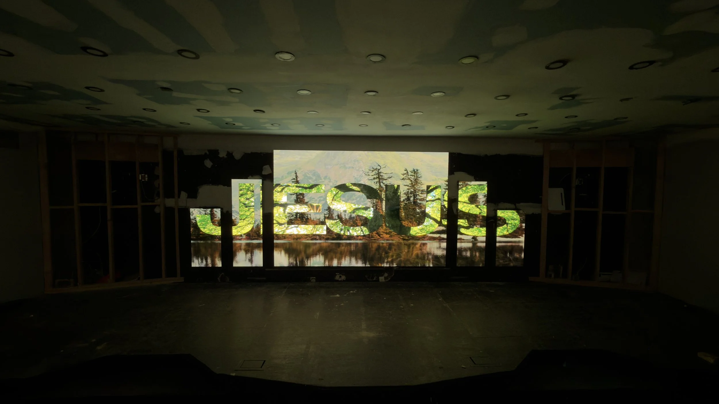 Indoor space under construction with framed walls and a large window displaying a nature scene with trees and mountains, the word 'JESUS' appears as a projected or printed overlay on the window.