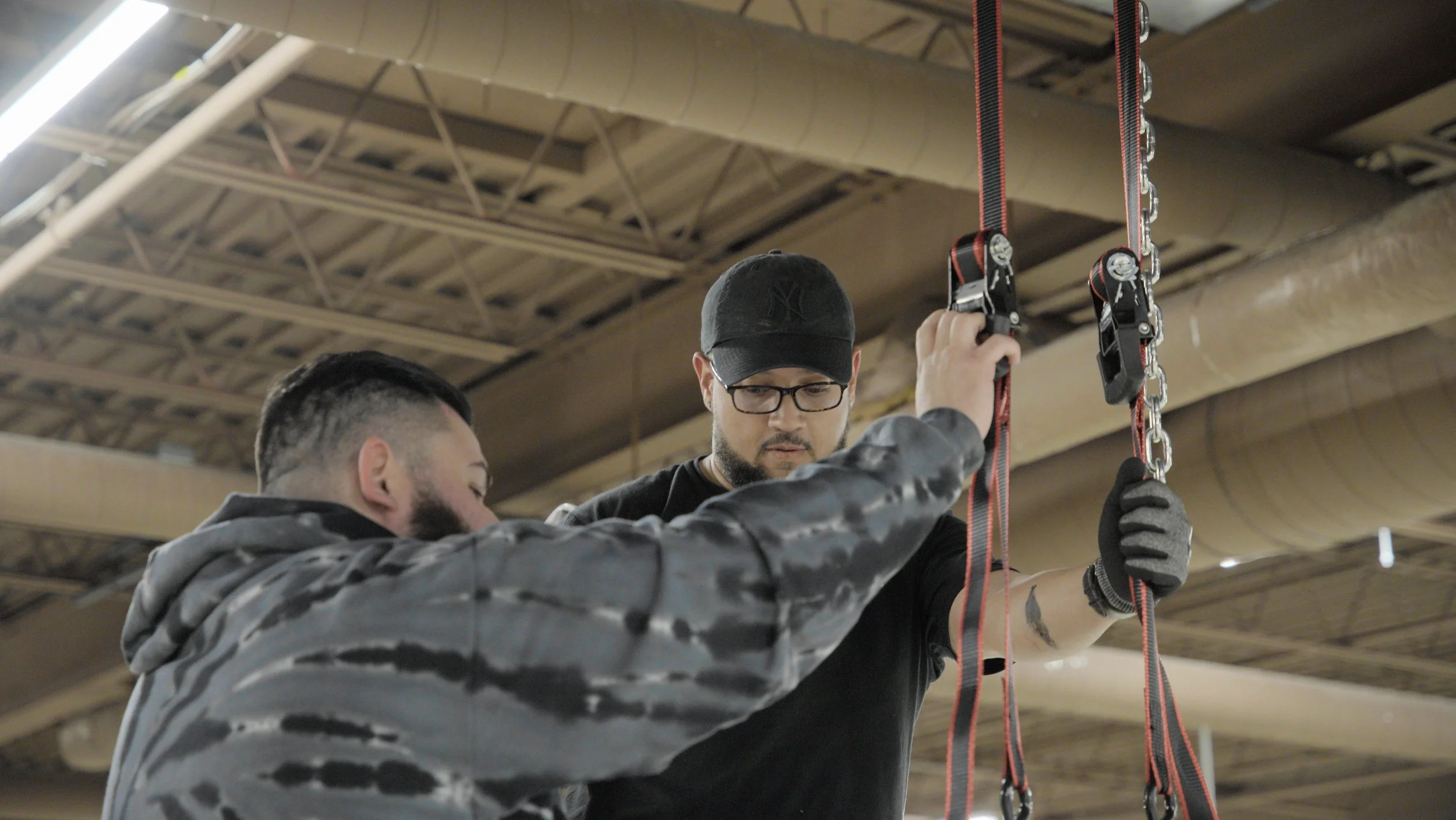 Two men working together in an industrial setting, with one holding a piece of equipment or apparatus attached to red straps with chains, possibly installing or repairing it under a high ceiling with exposed pipes and beams.