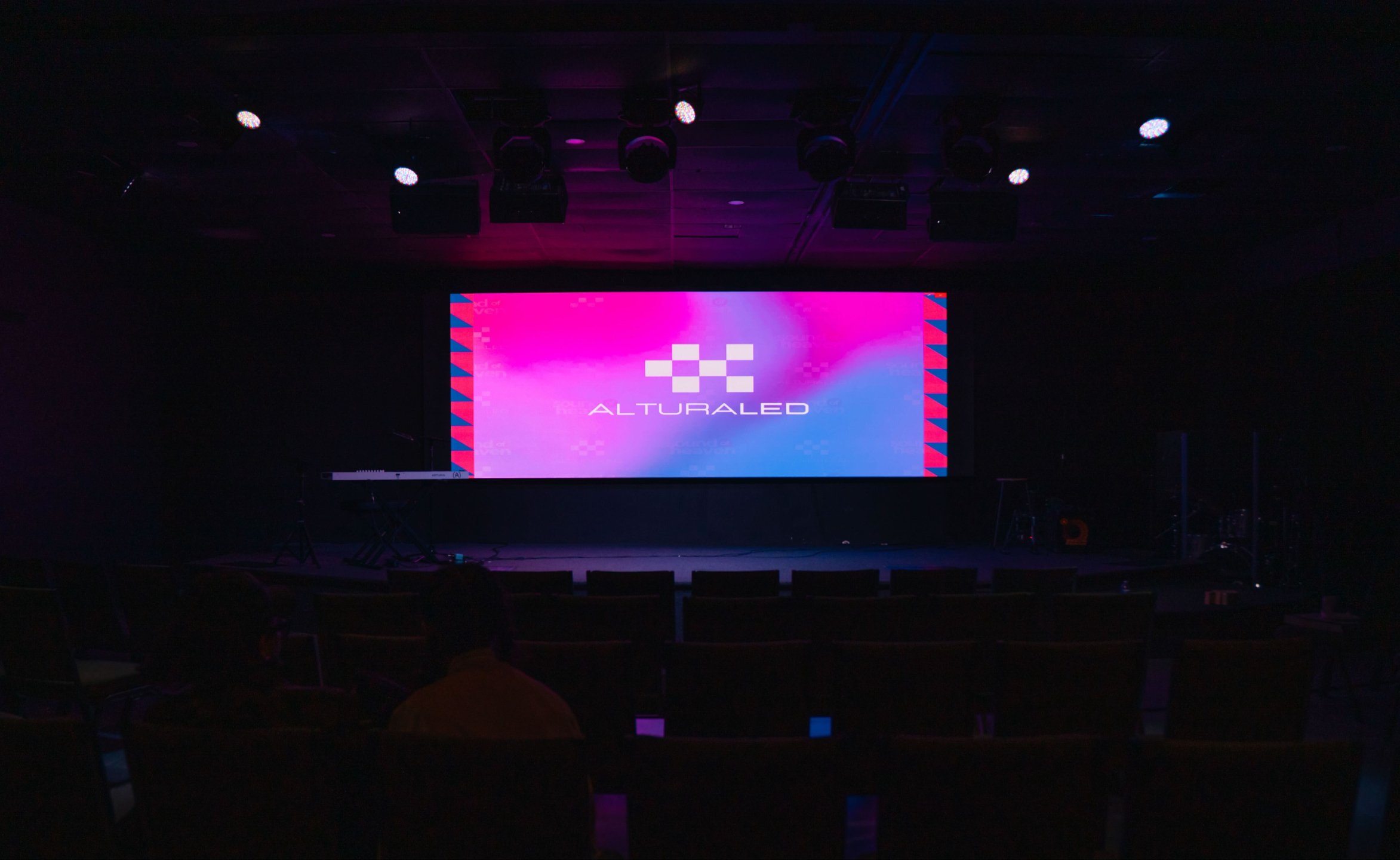 A dark auditorium with a large digital screen displaying the word 'ALTURALED' along with a pixelated logo, surrounded by stage lights.