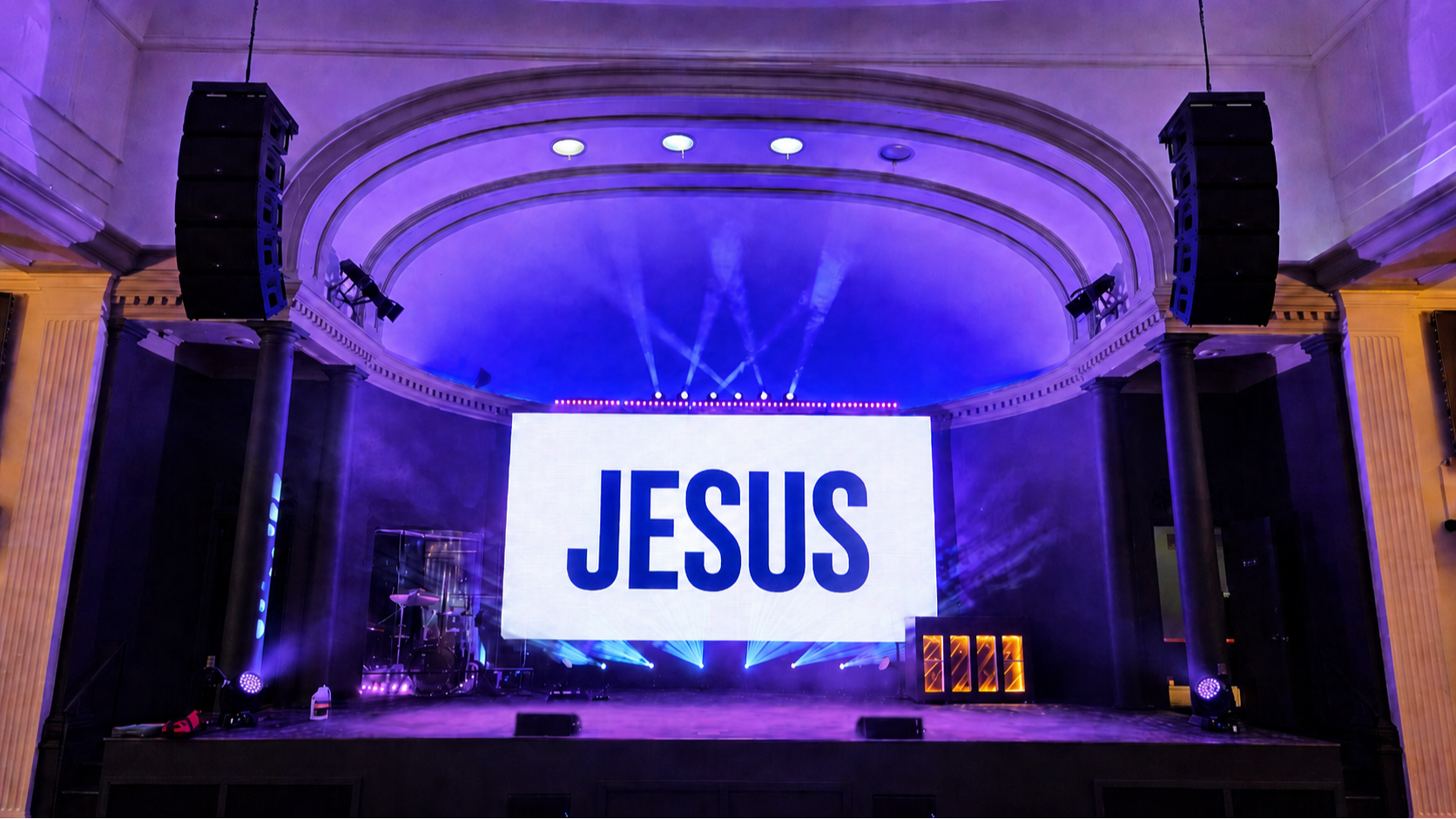 Stage with large screen displaying the word 'JESUS', surrounded by purple and blue lighting and large speakers on both sides.