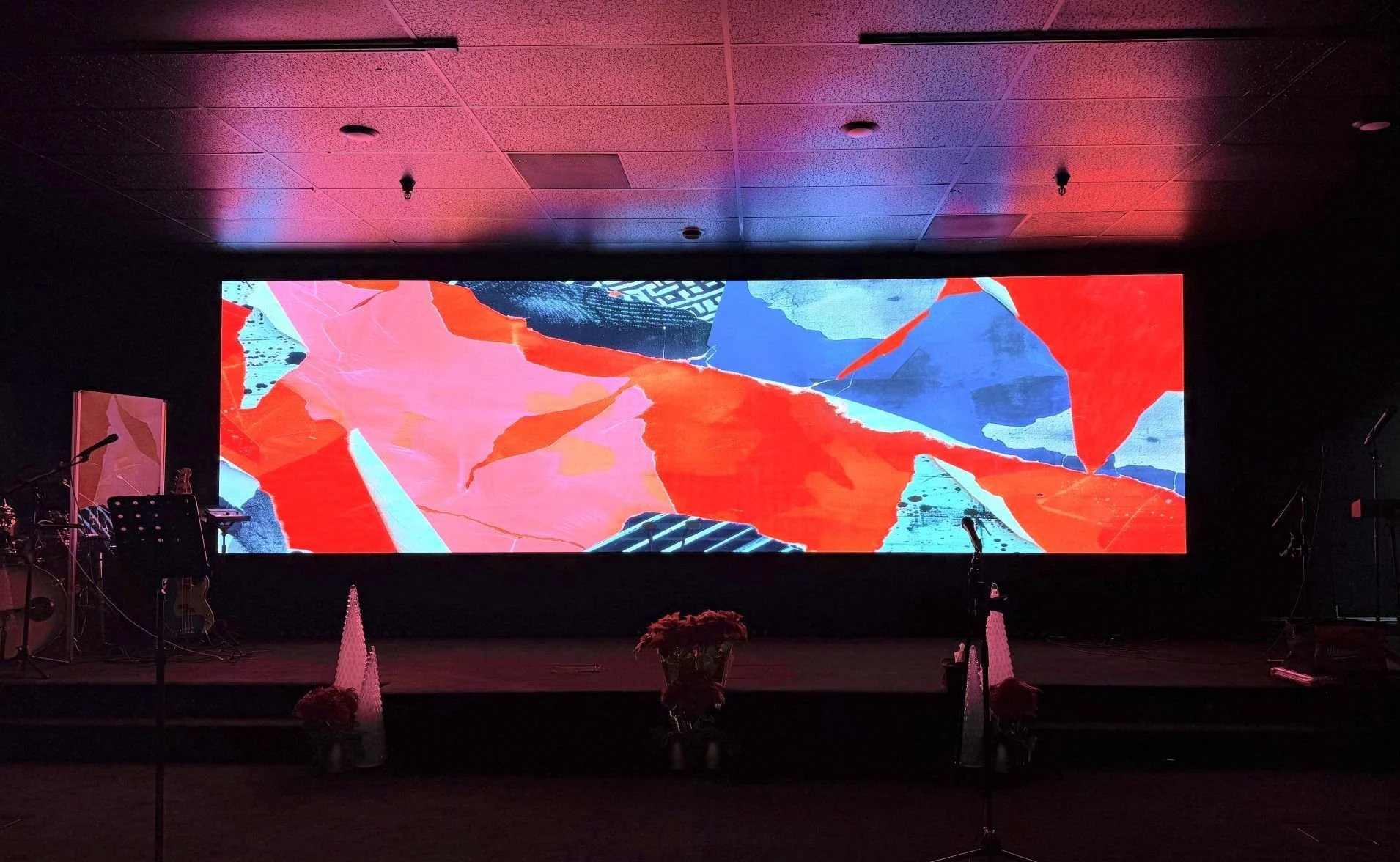 A large digital screen displaying an abstract art piece with red, blue, pink, and black shapes in a dark room with microphones, musical instruments, and floral arrangements in the foreground.