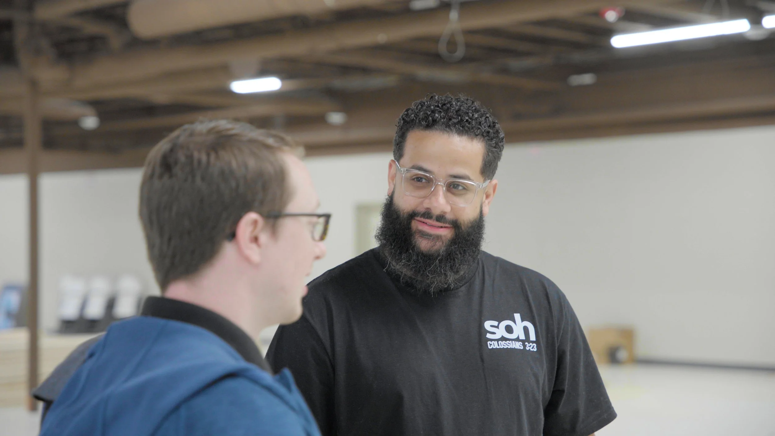Two men having a conversation indoors. The man on the right has curly hair and a beard and is wearing glasses and a black t-shirt with the words 'sah' and 'Colossians 3:23' printed on it. The man on the left has short, light brown hair and is wearing glasses and a blue hoodie.