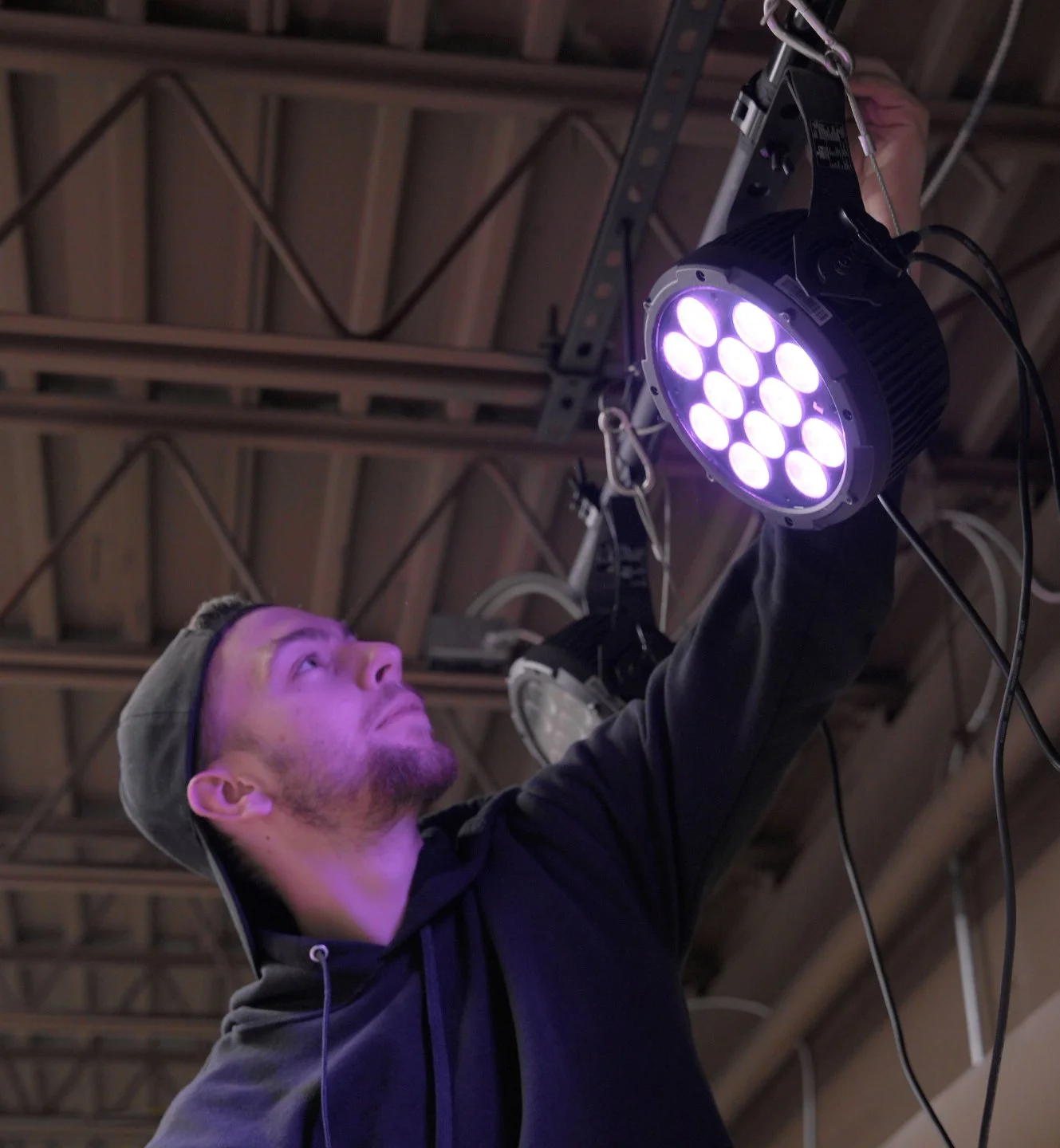A man adjusts or checks a large black stage light with multiple LED bulbs, hanging from the ceiling in an industrial or warehouse setting.