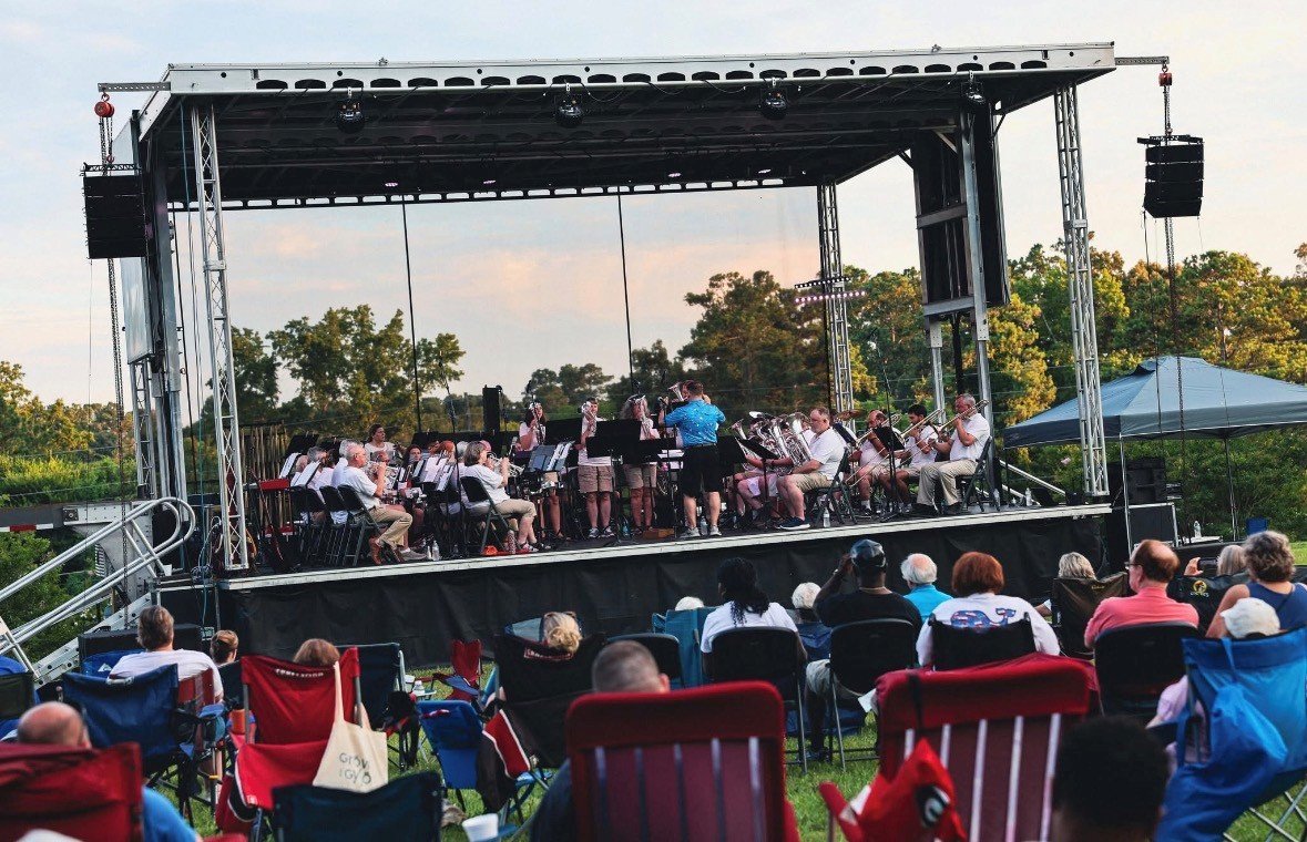 Roman Festival Brass Band  performing on an outdoor stage during a concert at sunset, with audience seated in lawn chairs facing the stage.