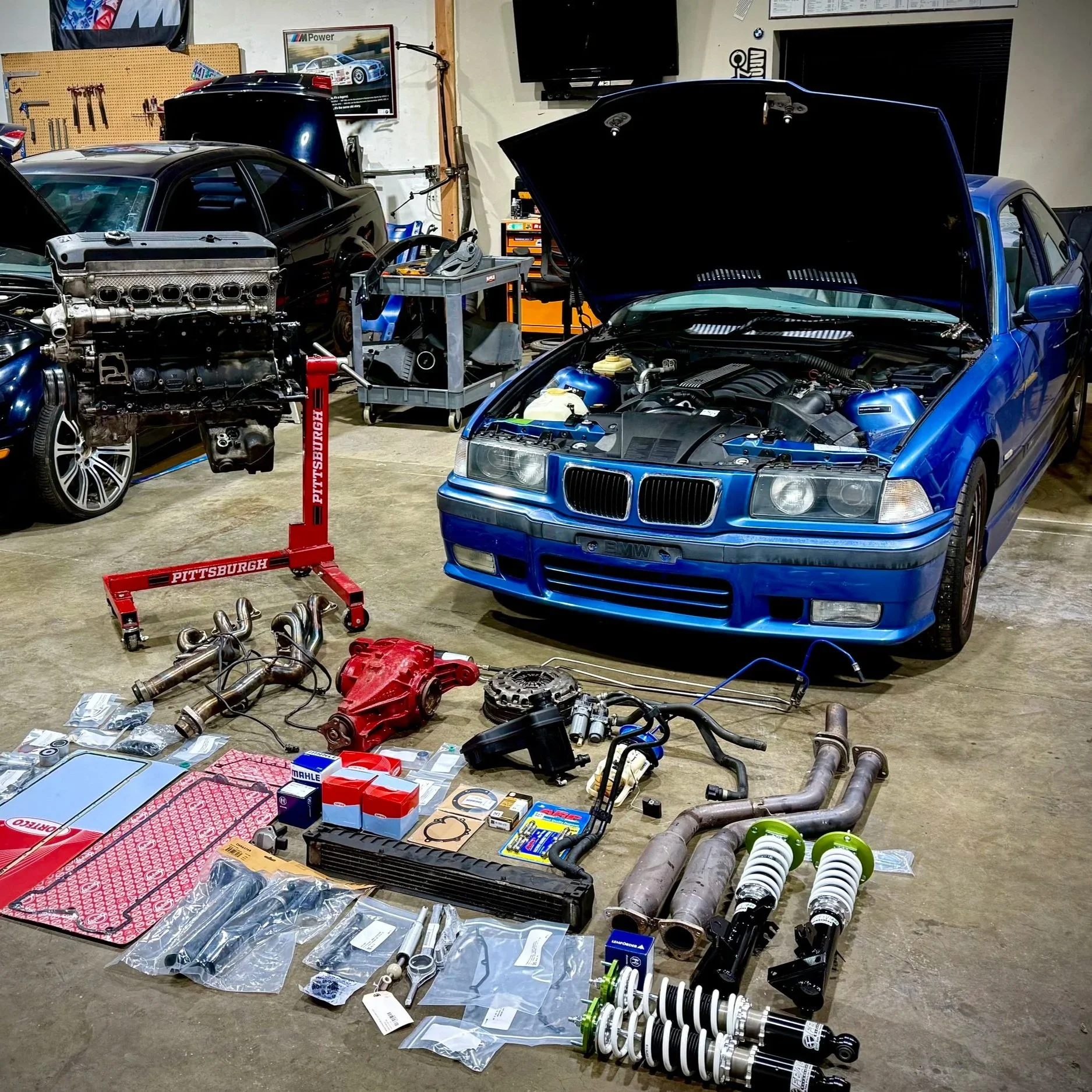 Garage workshop with disassembled car parts, tools, and automotive parts including a blue BMW with its hood open, a black car, various exhaust components, shocks, and boxes of parts laid out on the floor.