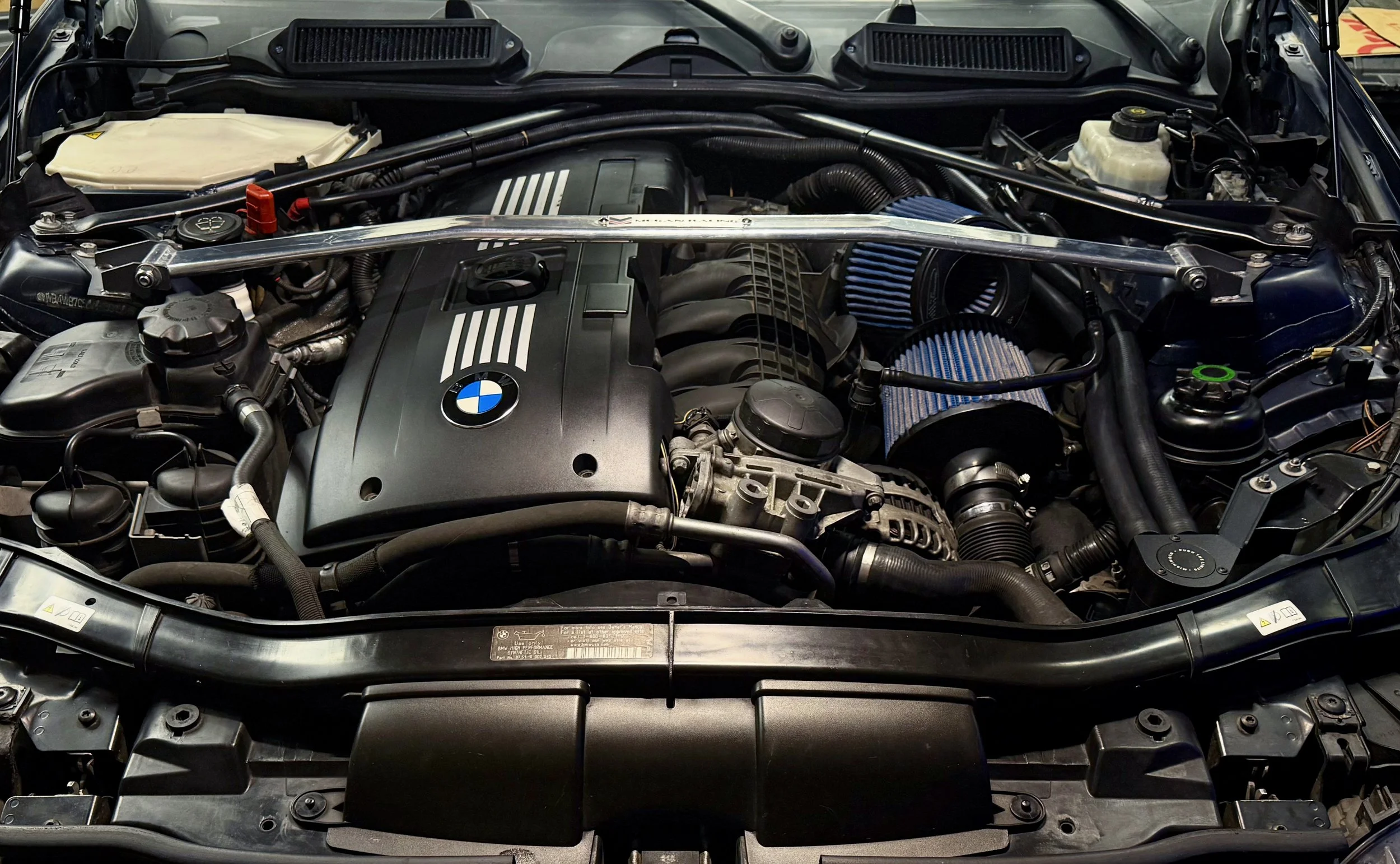 Close-up of a car engine bay, featuring a BMW engine, air filters, hoses, and various engine components.