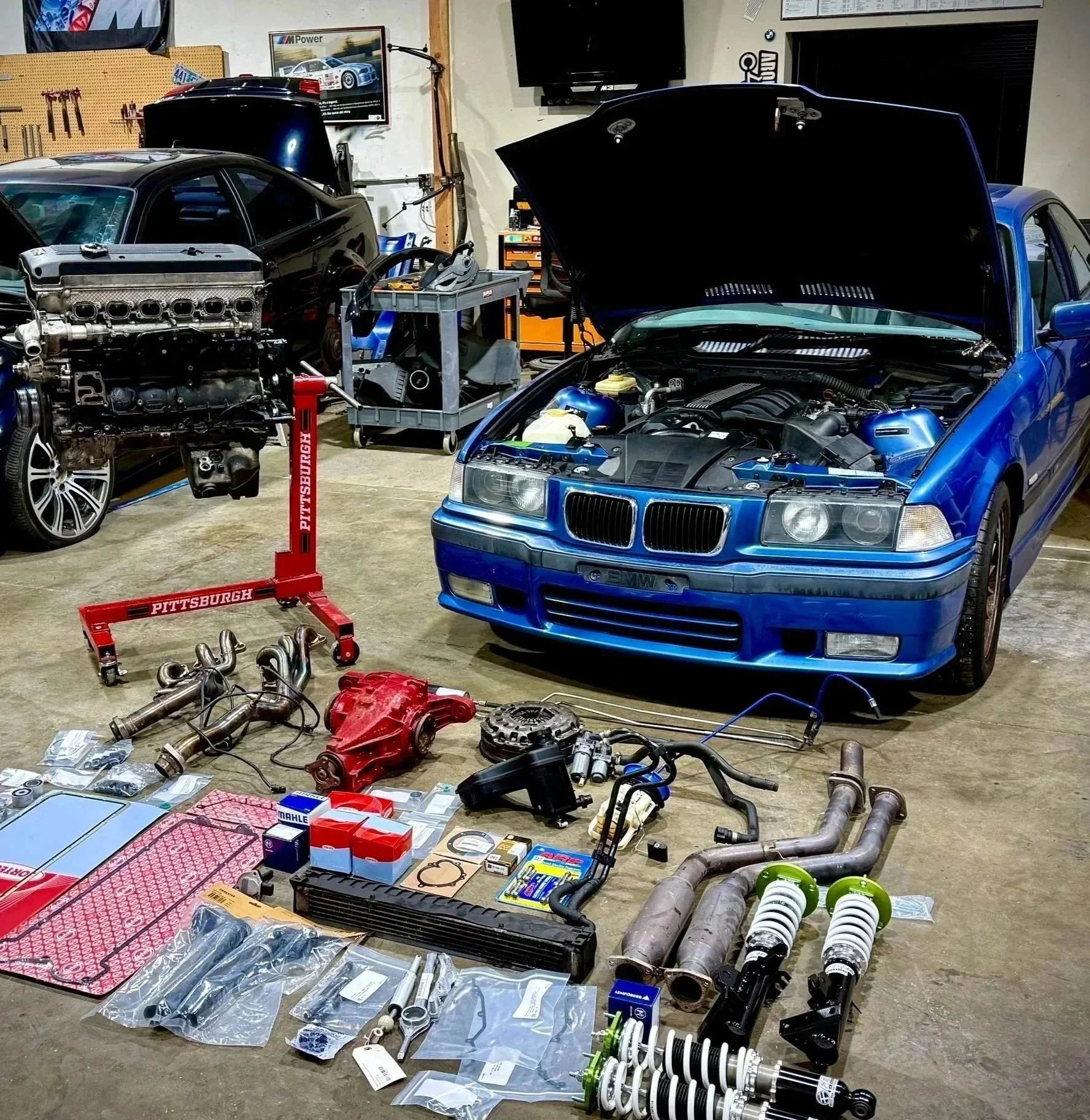Garage workspace with blue BMW car having open hood, various car parts, tools, and accessories scattered on the floor and workbenches, including an engine, exhaust components, shocks, and car-related boxes.