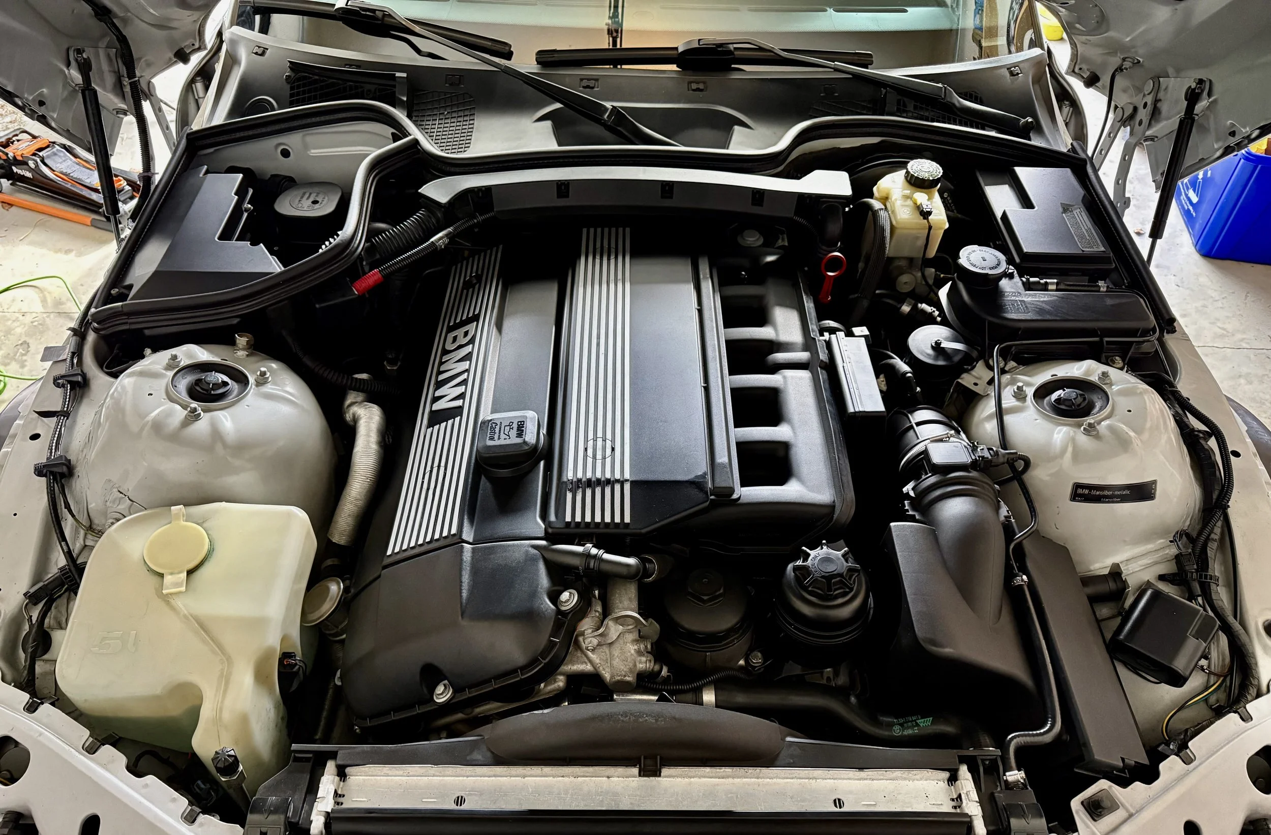 The image shows the engine bay of a BMW vehicle with the hood open, revealing the engine components, fluid reservoirs, and various hoses and wires.
