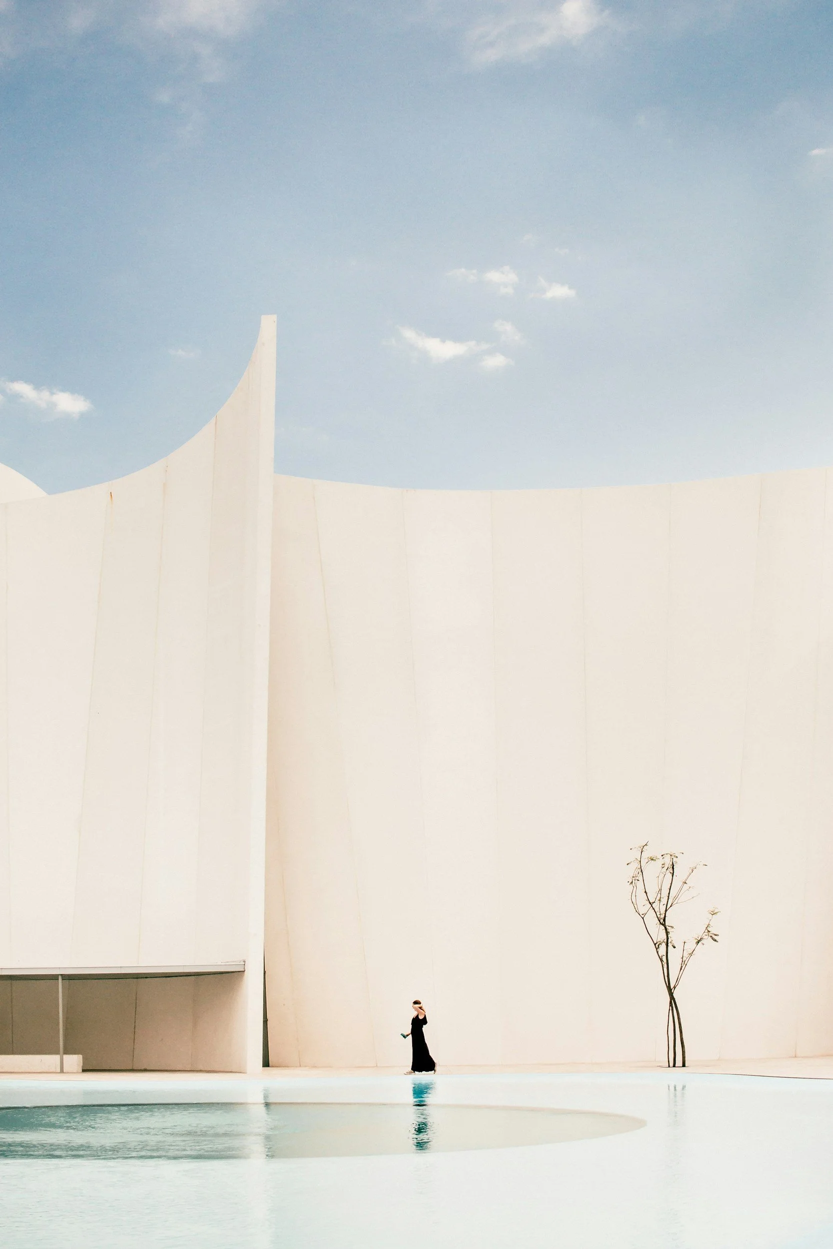 A woman in a long black dress standing near a reflecting pool in front of a modern white building with curved walls, a leafless tree, and a clear blue sky with a few clouds.