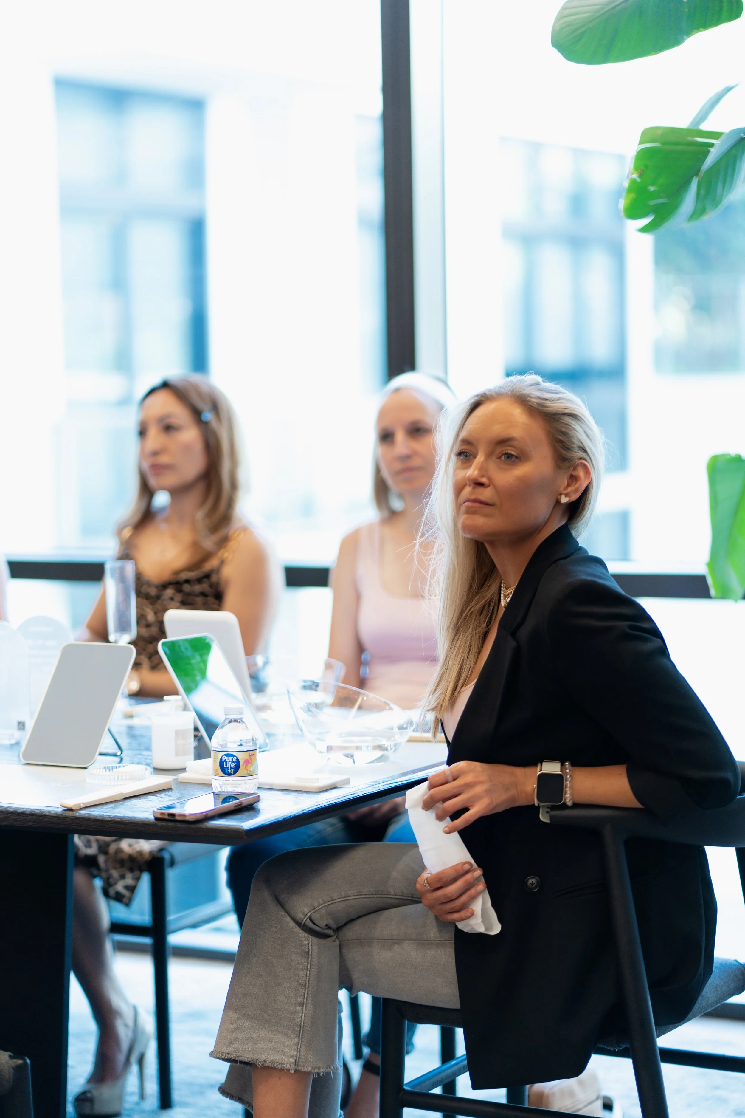 Four women in a meeting room with large windows, sitting at a table with phones, laptops, and meeting materials, engaged in a discussion.