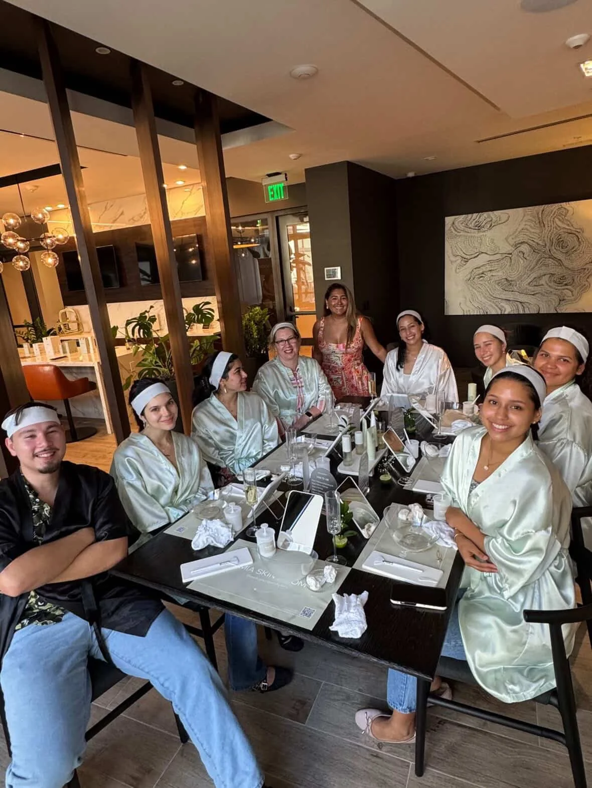 A group of women in matching satin robes and headbands sitting at a dining table in a restaurant, smiling for the camera.