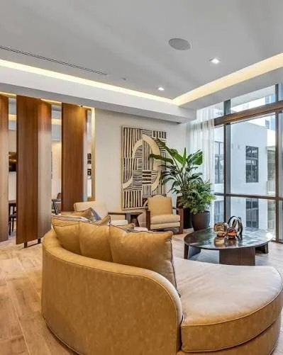 Modern living room with beige curved sofa, coffee table, large window, potted plants, and artwork on the wall.