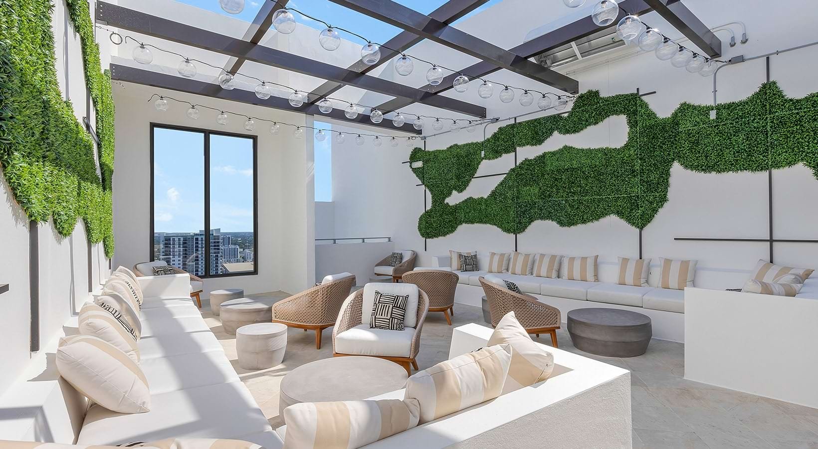 Indoor rooftop lounge with white walls, large window, string lights, green wall art, and modern furniture including sofas, armchairs, and stools.
