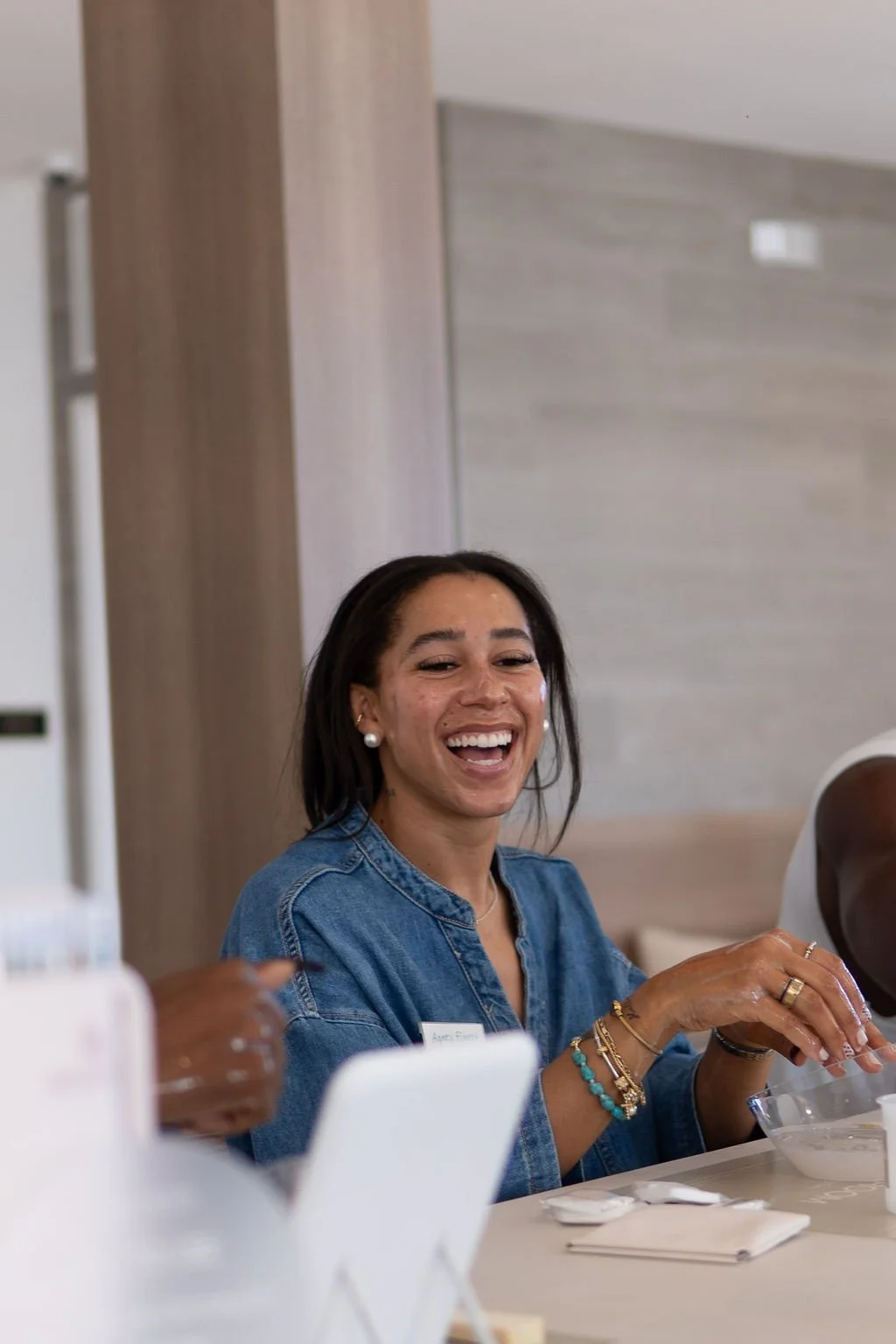 A woman with dark hair, wearing a denim jacket and pearl earrings, is smiling and sitting at a table during a gathering.