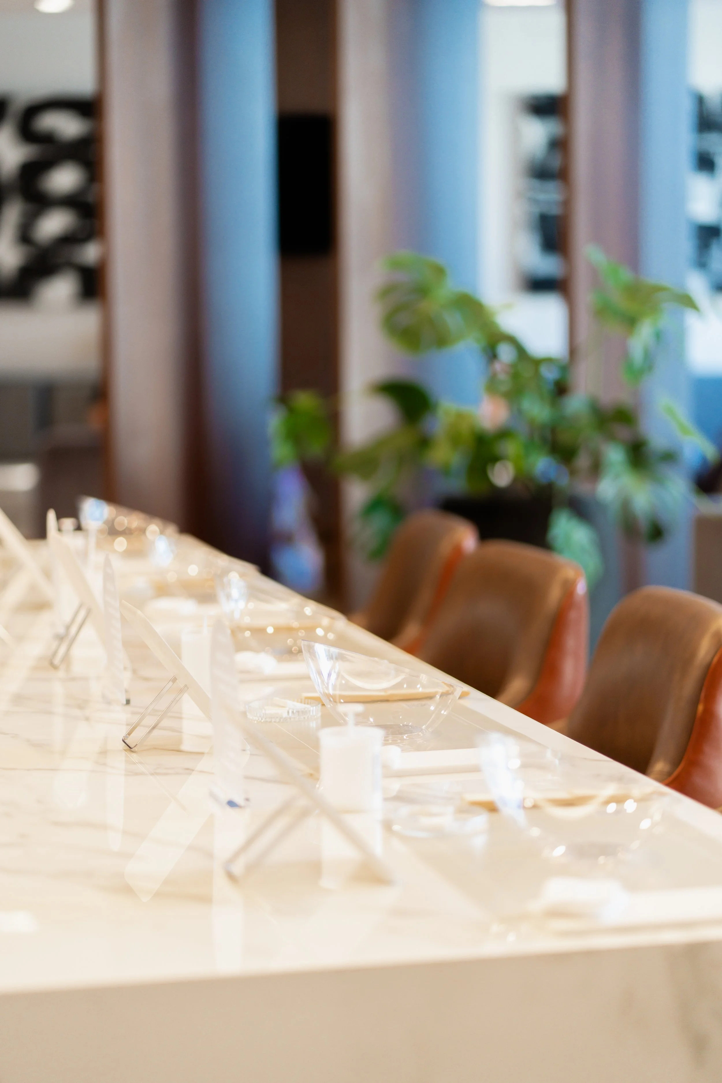 A restaurant or banquet room setup with a long white table, transparent bowls, and utensils. Behind the table are brown leather chairs and a large plant. In the background, there are vertical wooden beams and windows.