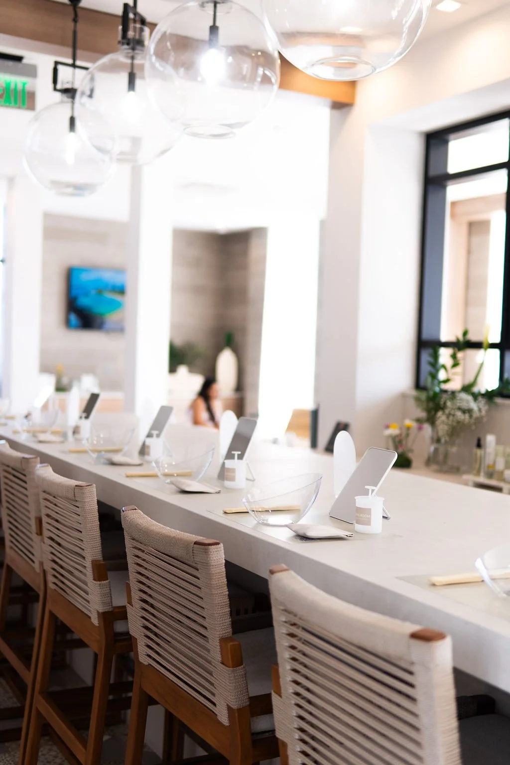 A modern restaurant interior with a long white table set with bowls, snack containers, and name cards, accompanied by wooden chairs with woven backs, and decorative lighting fixtures hanging from the ceiling.