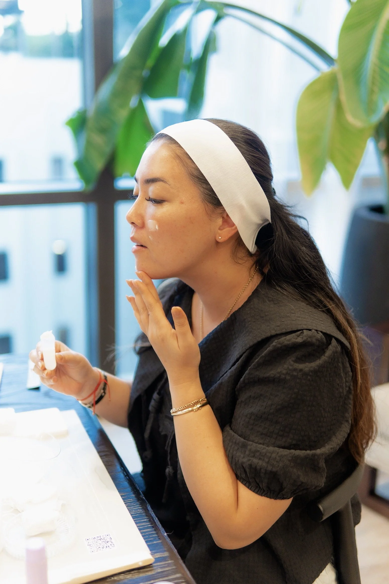 A woman with dark hair, wearing a white headband and black top, is sitting at a table with skincare or cosmetic products, applying or examining her face.