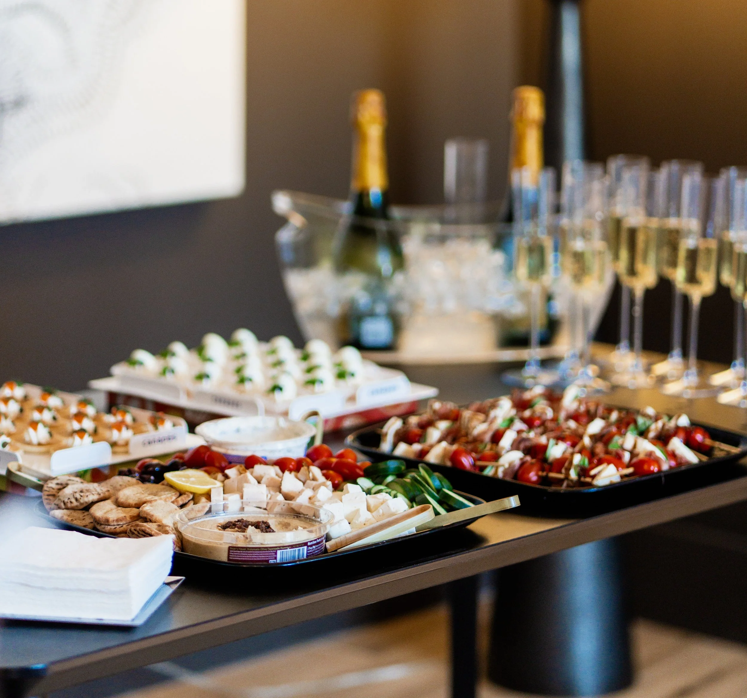 A buffet table displaying various types of desserts and snacks, including small pastry bites, cookies, and a platter of fruit, with bottles of beverage behind the table.