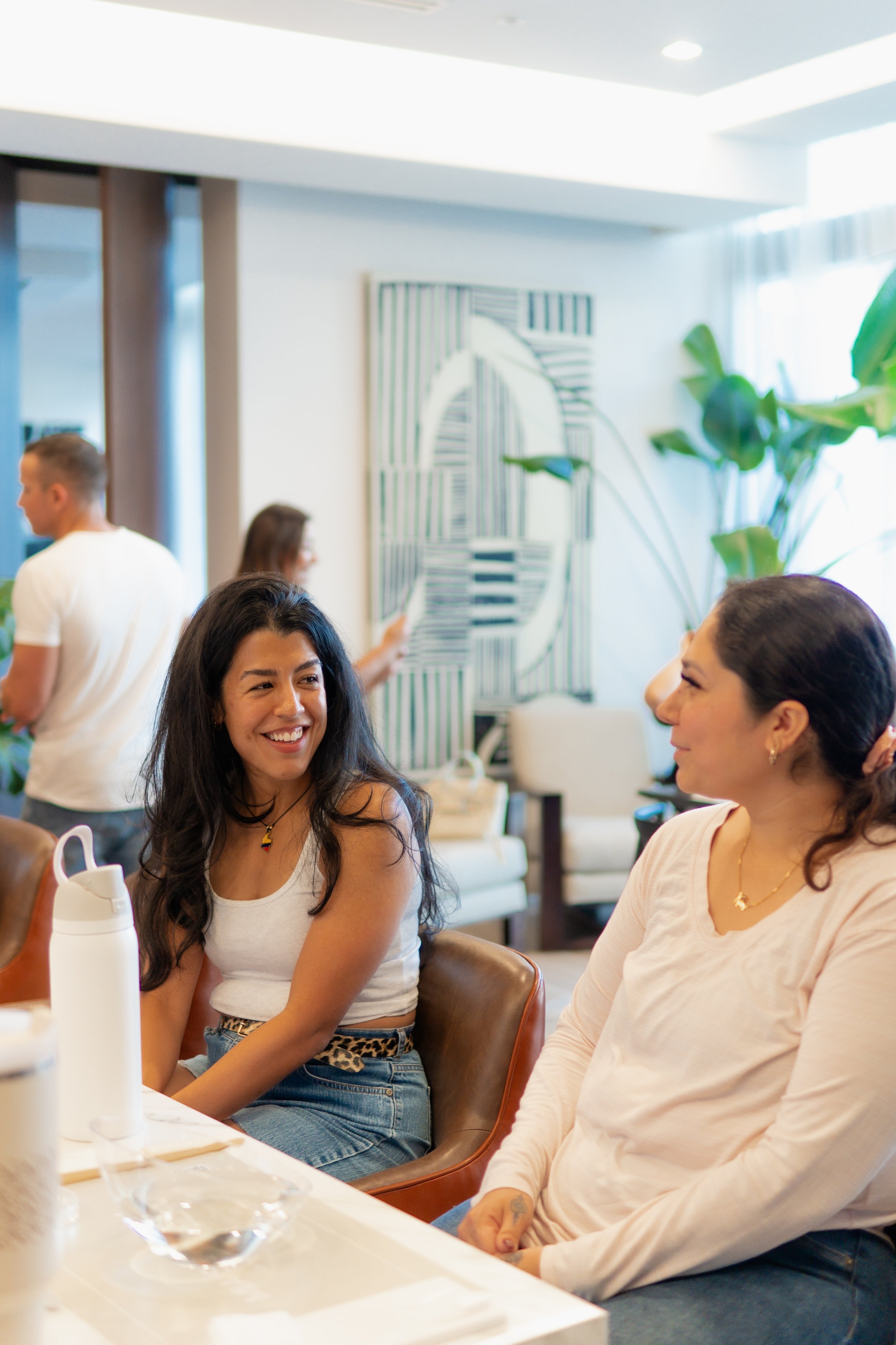 Two women seated at a table smiling and engaging in conversation in a bright room with green plants and abstract artwork in the background.