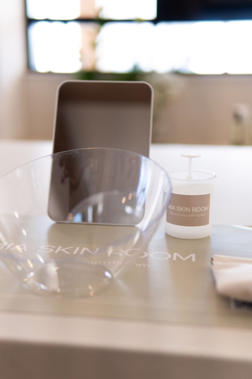 A skincare product, a mirror, and a glass bowl on a table in a bright room with a window in the background.
