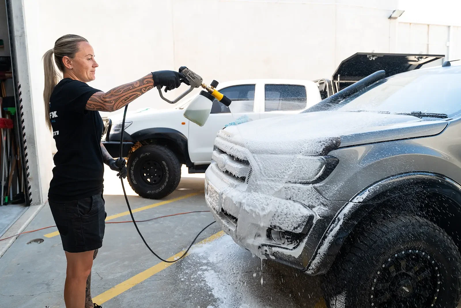 A woman pressure washing the front of a gray vehicle covered in soap foam in a parking lot, with a white truck and a garage door in the background.