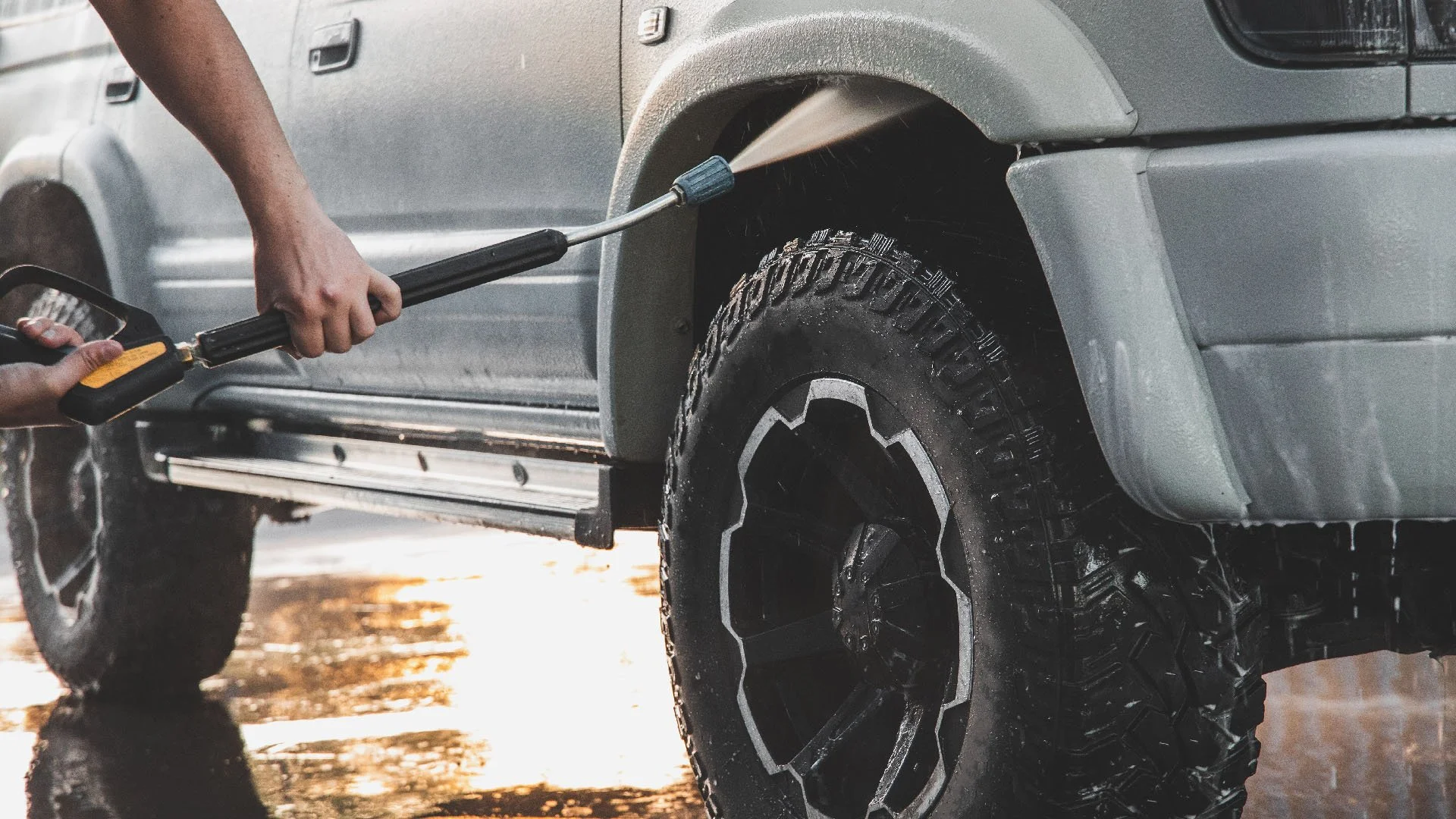 A person cleaning the tire of a gray SUV with a pressure washer.