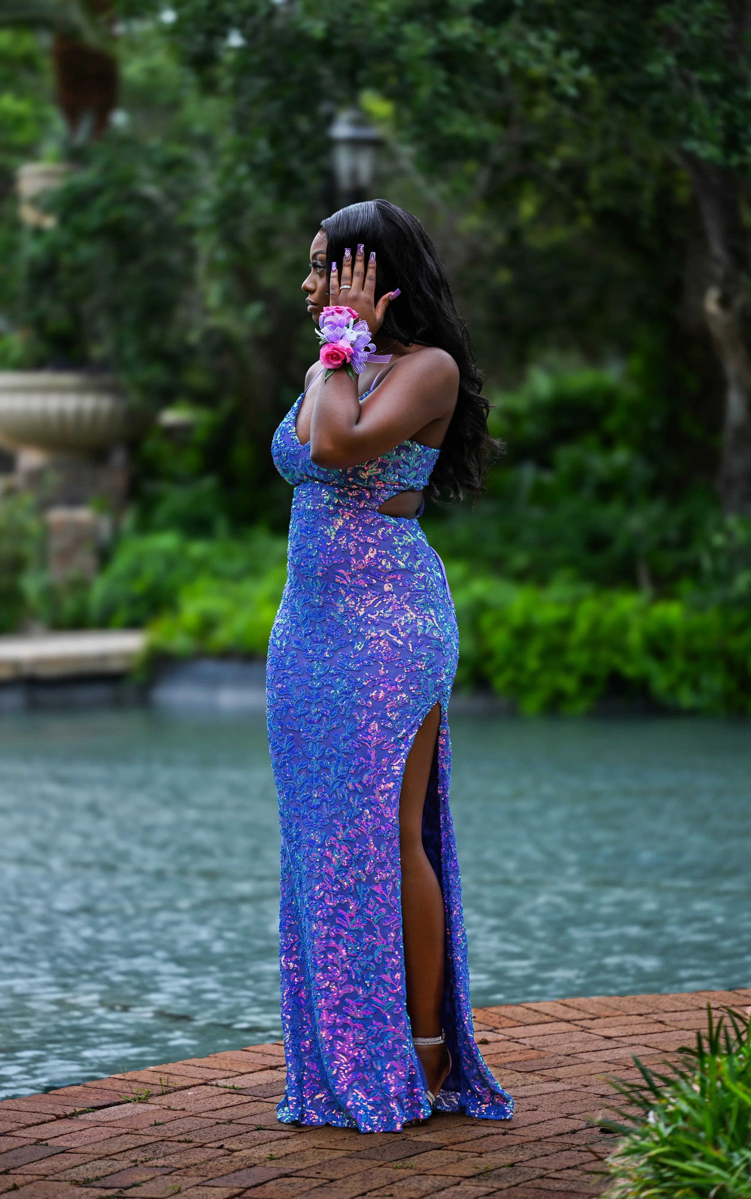 A woman in a purple and blue patterned formal gown standing on a brick pathway near a body of water, with lush green trees in the background. She is posing with her hand on her head and has floral wrist corsages.