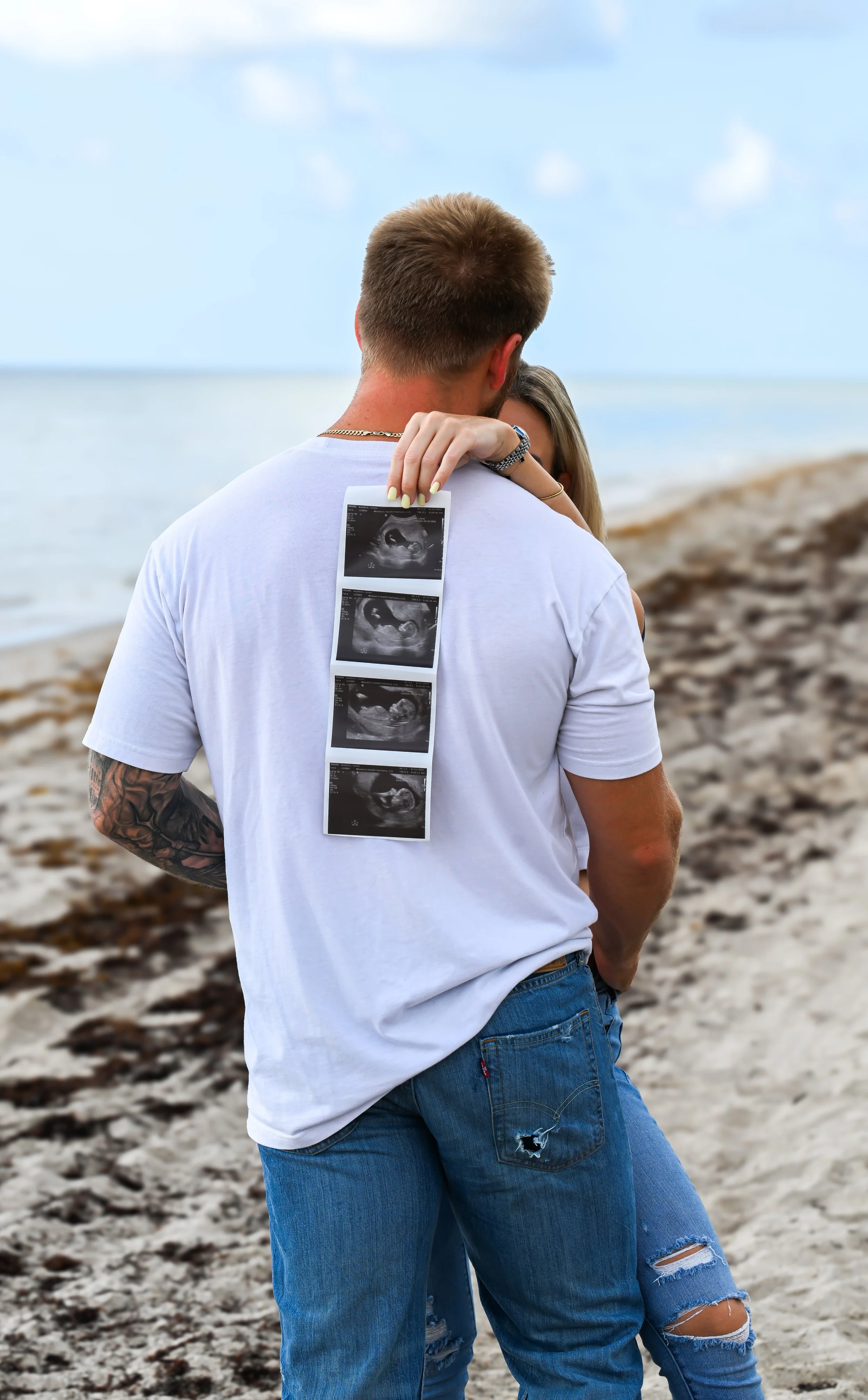 A man and woman on a beach, with the woman holding ultrasound images. The man has his back to the camera, and the woman is hugging him from the front.