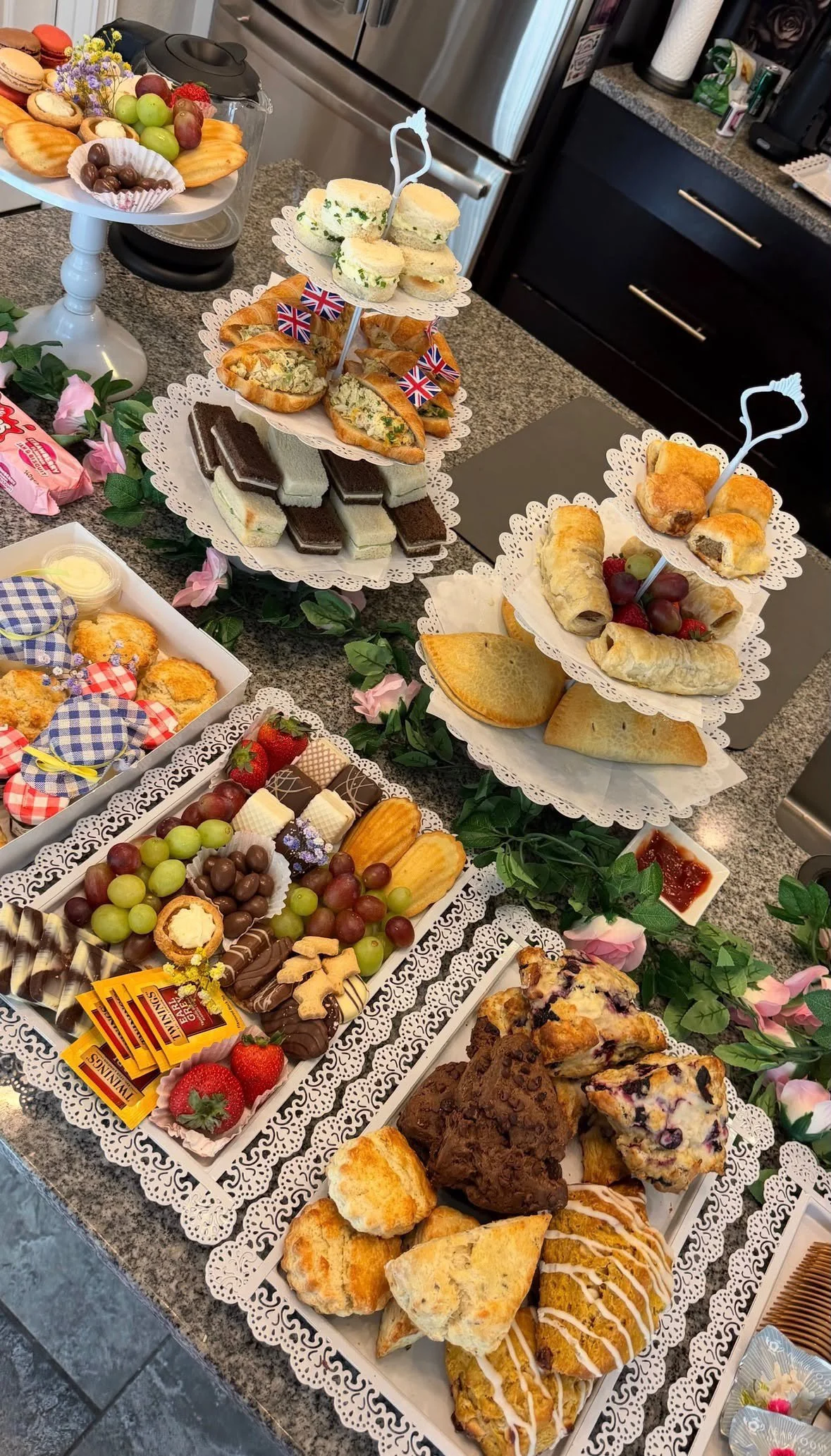 A dessert and snack display featuring fruit, sandwiches, pastries, cookies, and sweets arranged on tiered serving trays with decorative doilies, surrounded by artificial pink flowers.