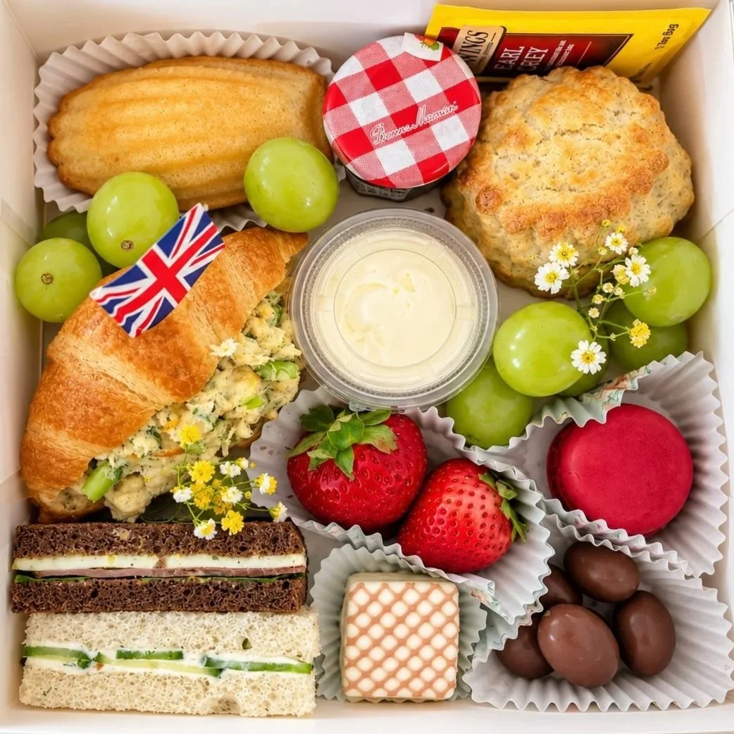 A box of assorted food including green grapes, strawberries, chocolate eggs, a creamed chicken salad croissant sandwich, a small cake, a scone, a jam jar, a small layered cake, cucumber sandwiches, and a container of deviled eggs, decorated with small flowers.