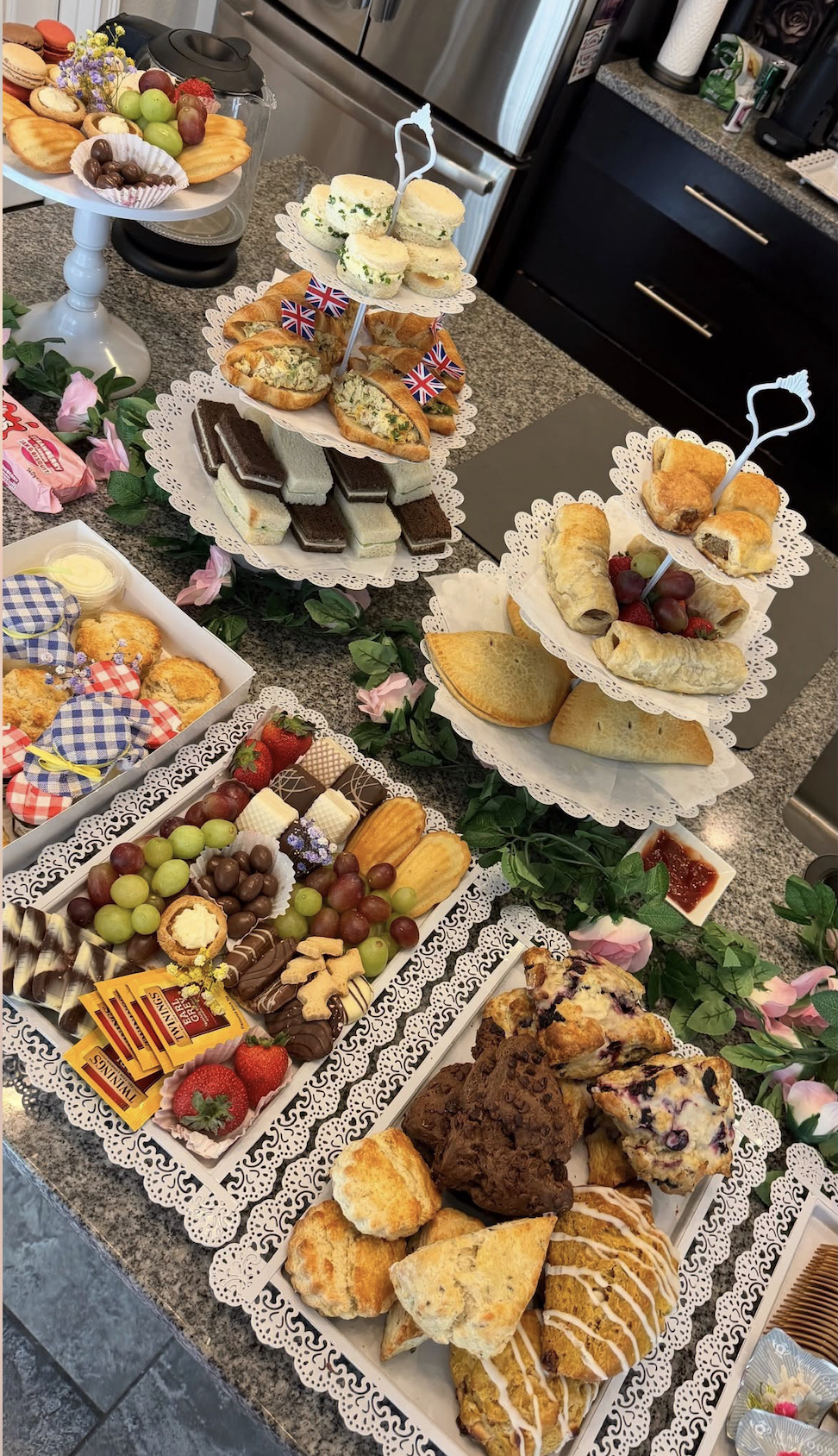 A variety of desserts and snacks arranged on lace and paper doilies, including pastries, cookies, chocolates, fruit, and mini sandwiches, decorated with small Union Jack flags, on a kitchen countertop.
