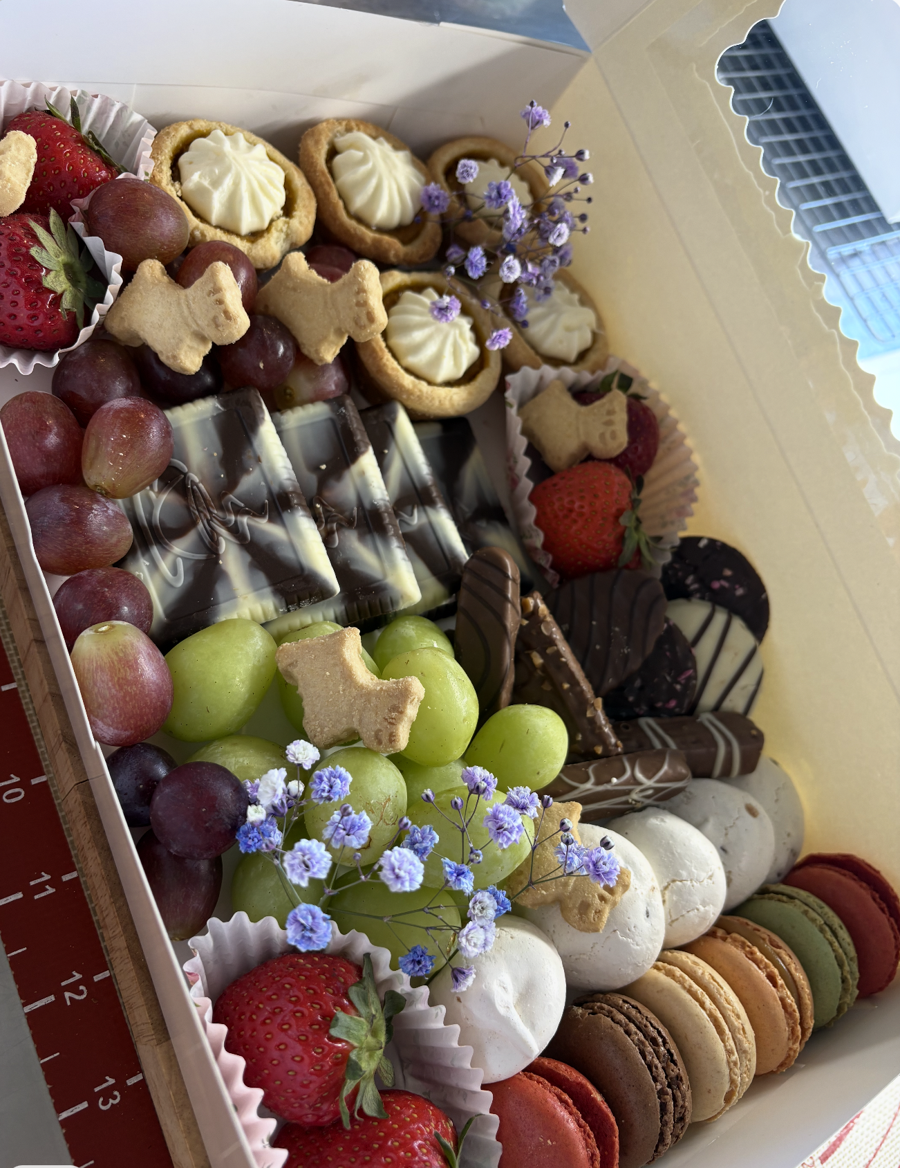 A box filled with various desserts and snacks including strawberries, grapes, cookies with white frosting, chocolates, and colorful macarons, some garnished with purple flowers.