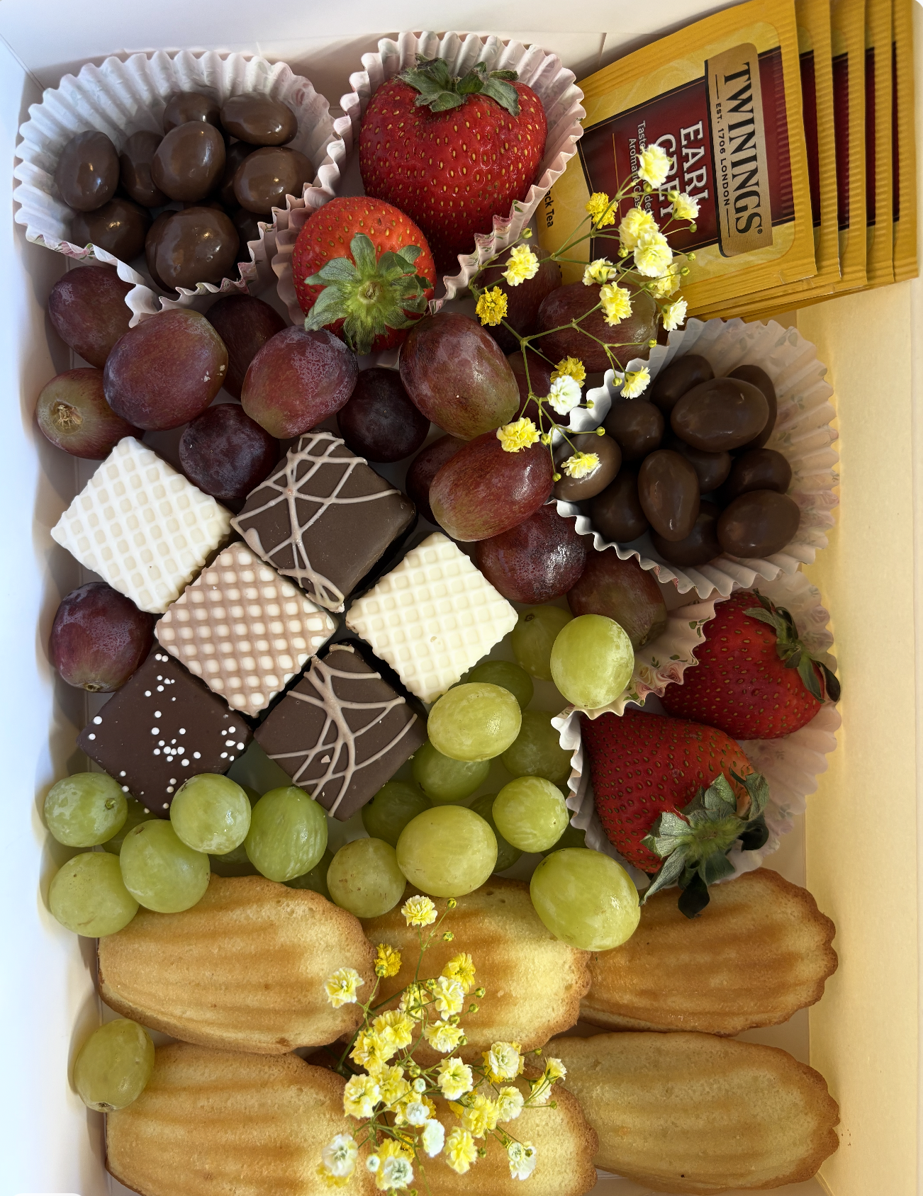 A box of assorted chocolates, fresh strawberries, green and red grapes, and cookies, with yellow flowers for decoration.