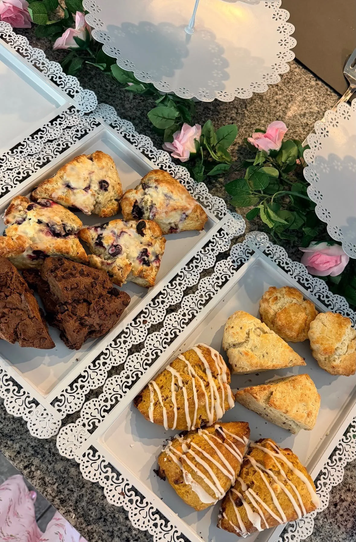 Assorted baked goods on white lace-edged trays, with pink roses and green leaves as decoration, on a granite countertop.