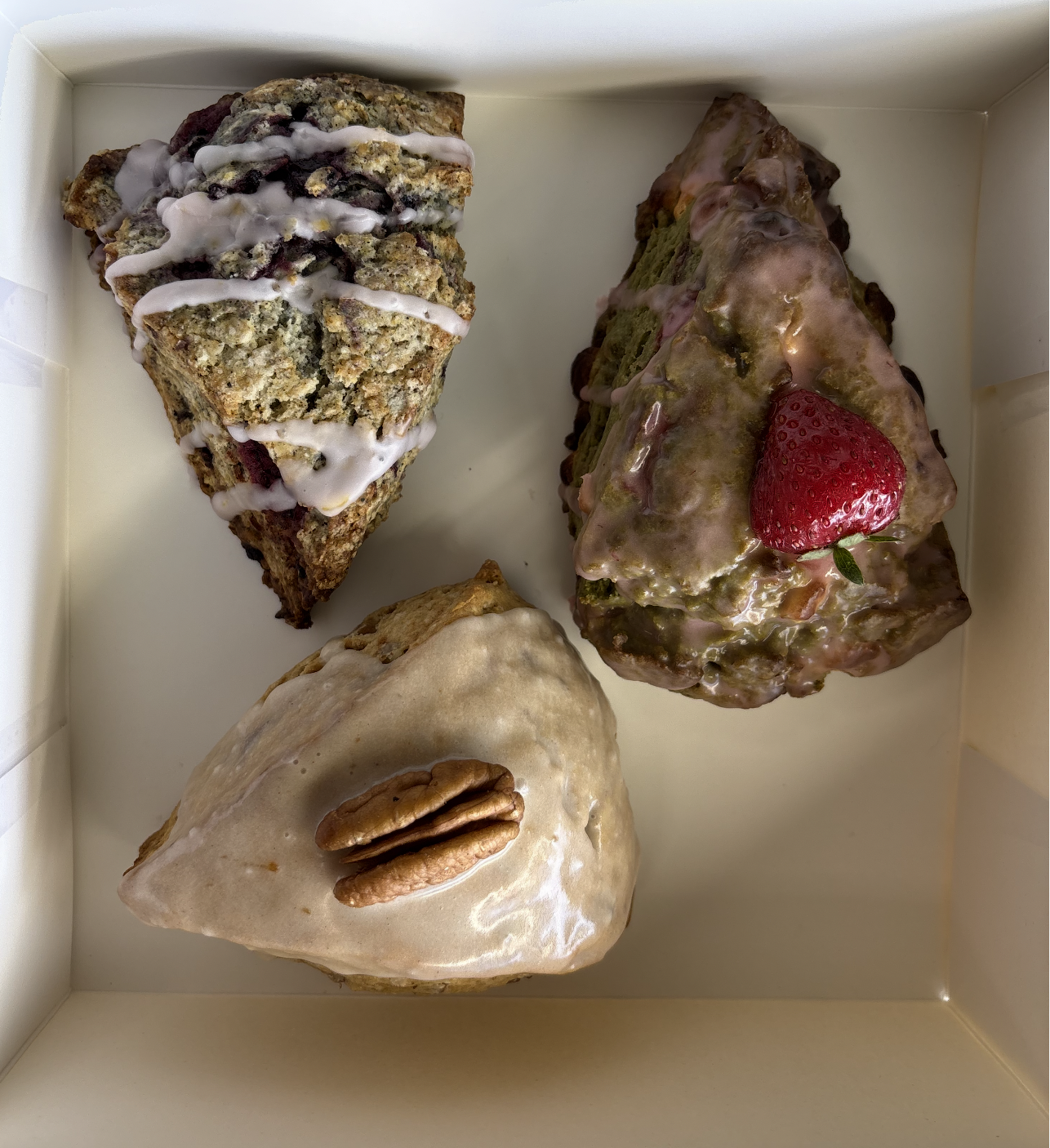 Three different slices of cake in a white box, one with nuts and icing, another with berries and a strawberry on top, and the third with a pecan nut.