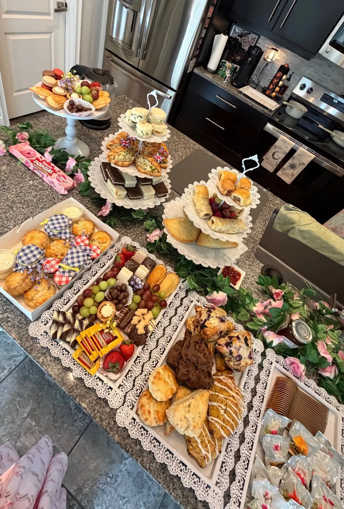 A large assortment of baked goods and snacks arranged on a kitchen table decorated with pink flowers and green leaves, including cookies, scones, pastries, cakes, chocolates, and fresh fruit.