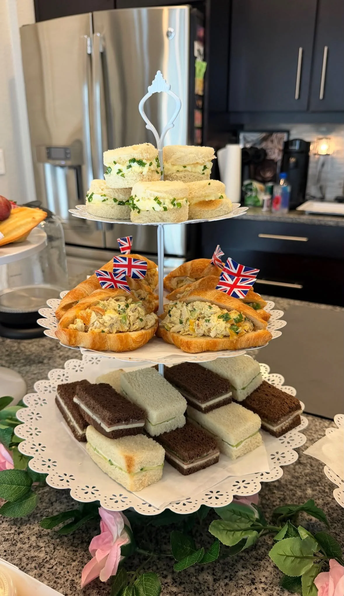 A four-tiered tray holds assorted finger sandwiches, mini cakes, and pastries, decorated with small Union Jack flags. The tray is set on a countertop with pink flowers and green leaves around it in a kitchen setting.