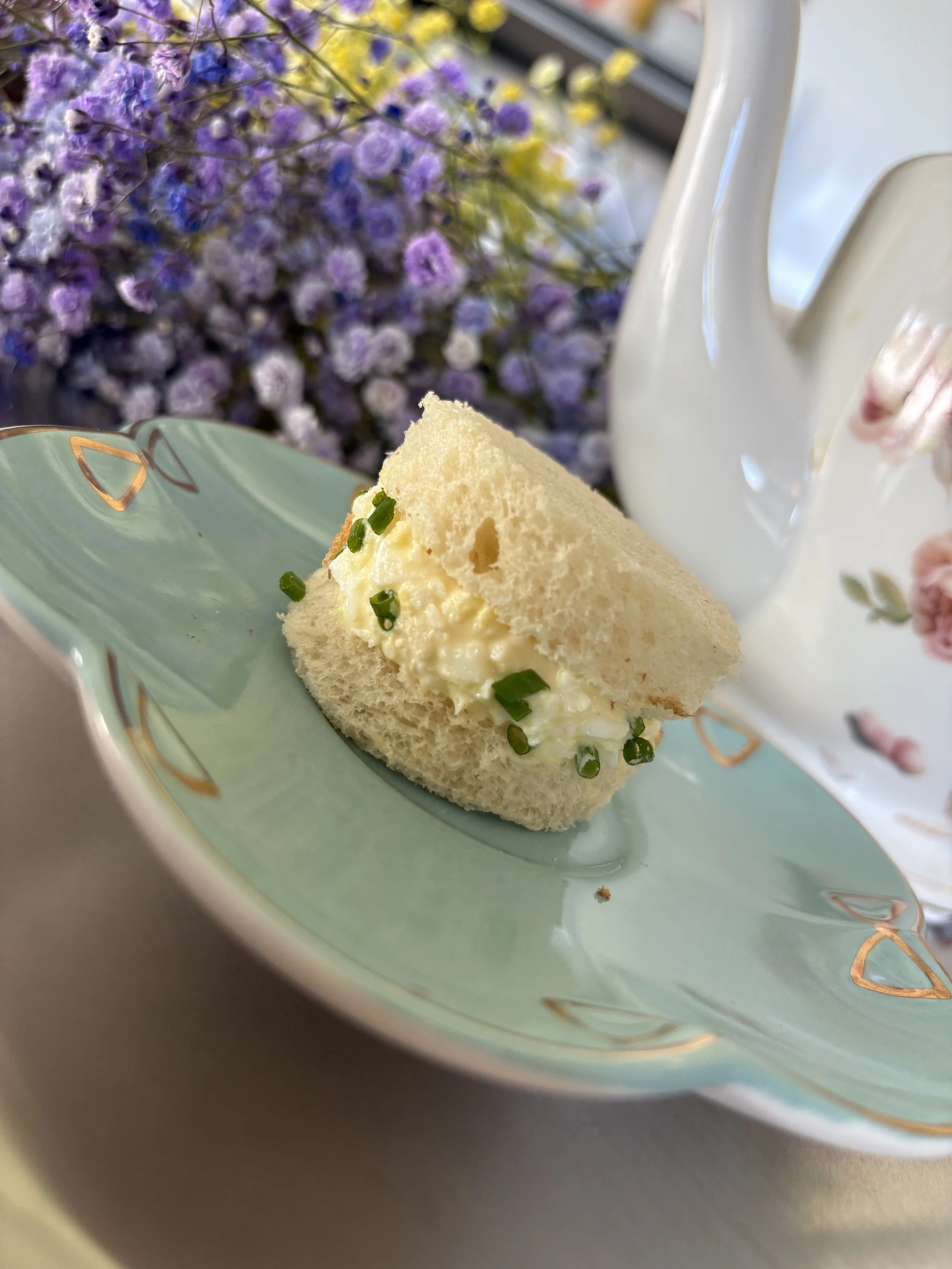 A slice of egg salad sandwich with chopped green onions in a light green dish with a floral pattern, placed on a table with a bouquet of purple and yellow flowers partially visible in the background.