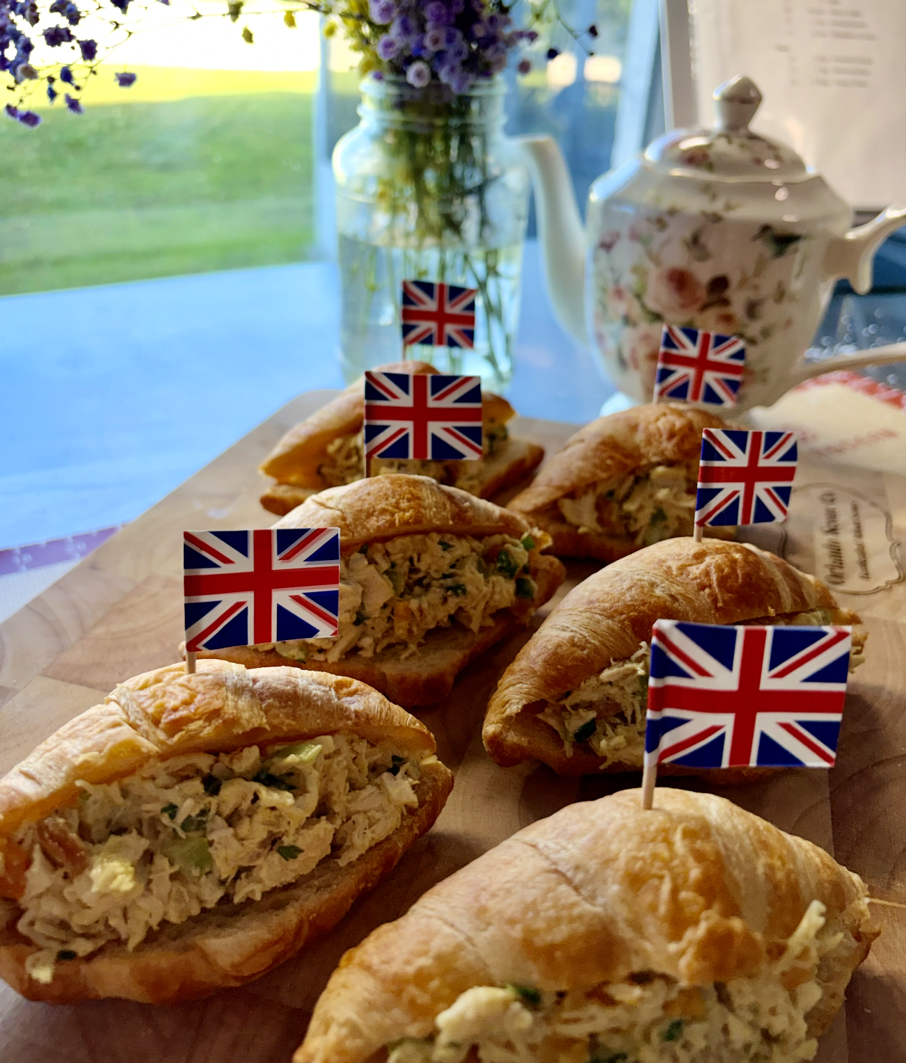 A tray of chicken salad croissants topped with small British flags, with a vase of purple flowers and a floral teapot in the background.